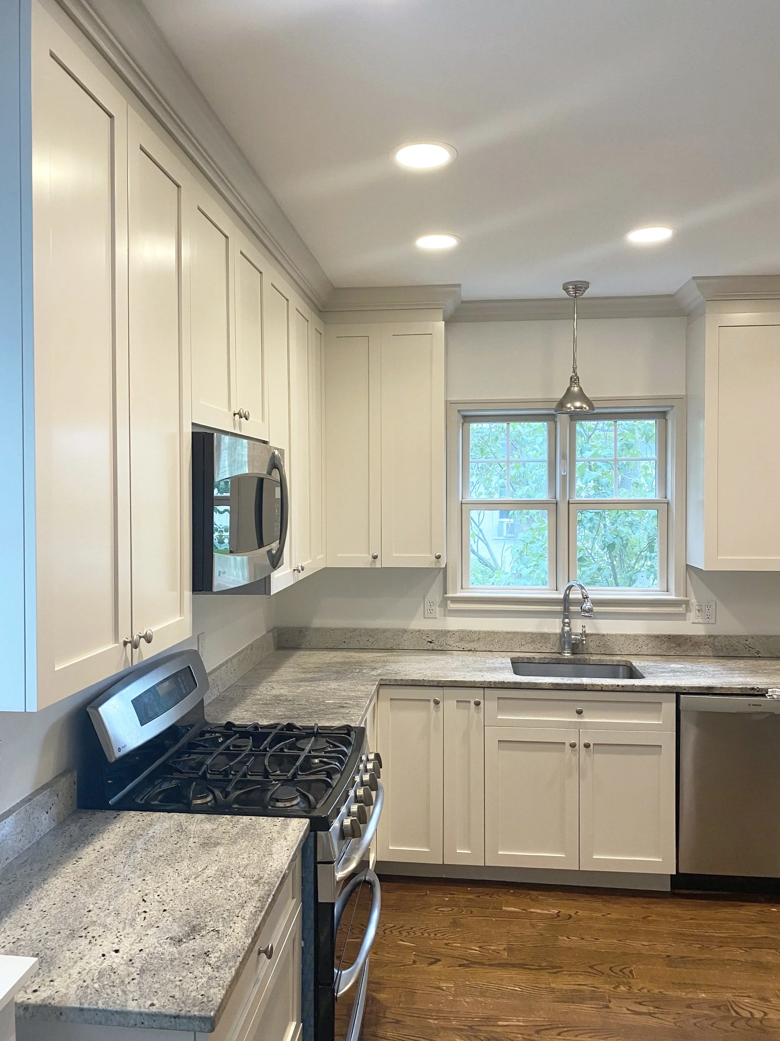 A modern kitchen with white cabinets, granite countertops, a stainless steel oven, microwave, and dishwasher. There is a window above the sink with greenery outside and ceiling lights overhead.