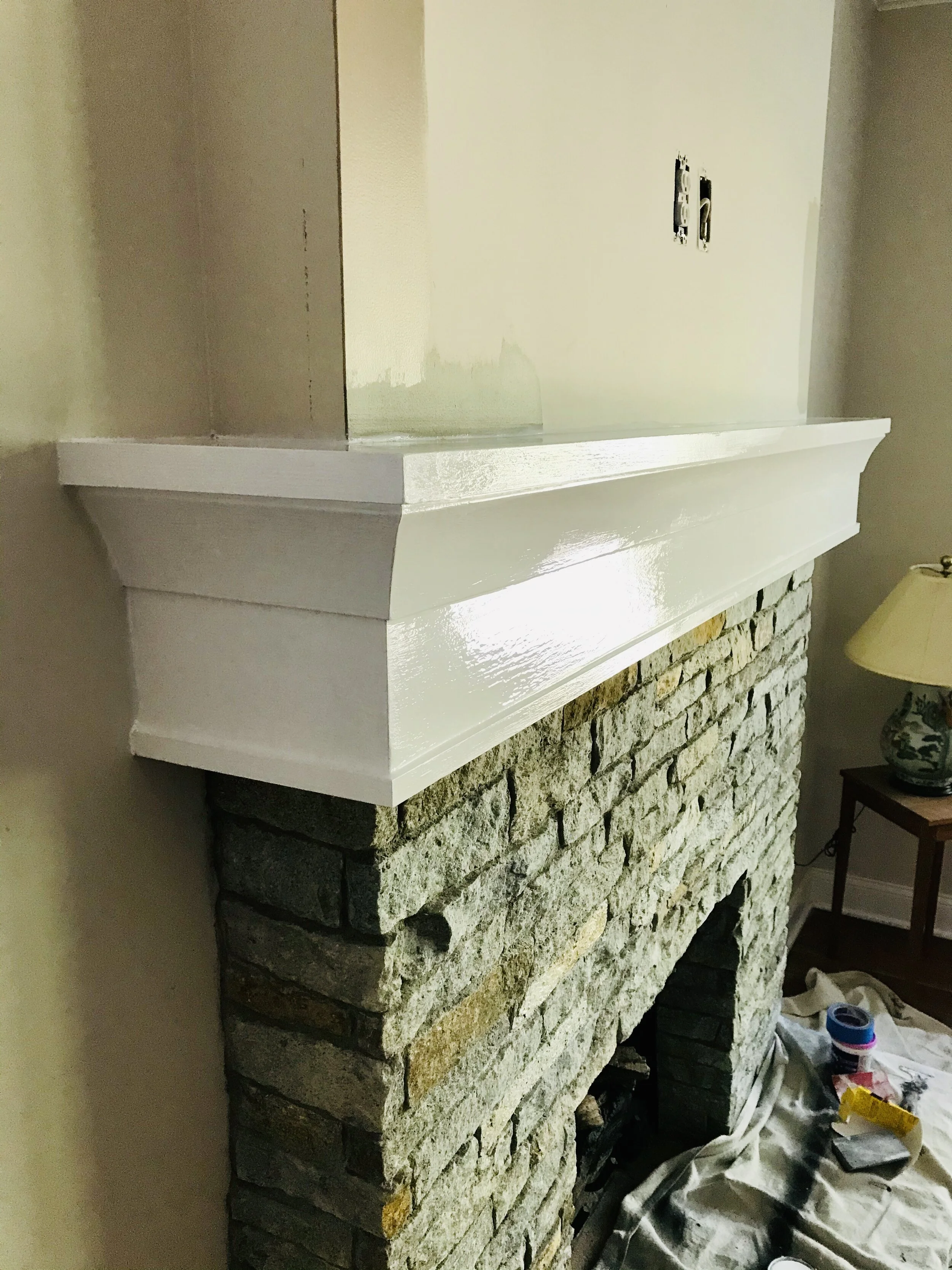 Close-up of a brick fireplace with a newly painted white mantel, with painting supplies on the floor nearby.