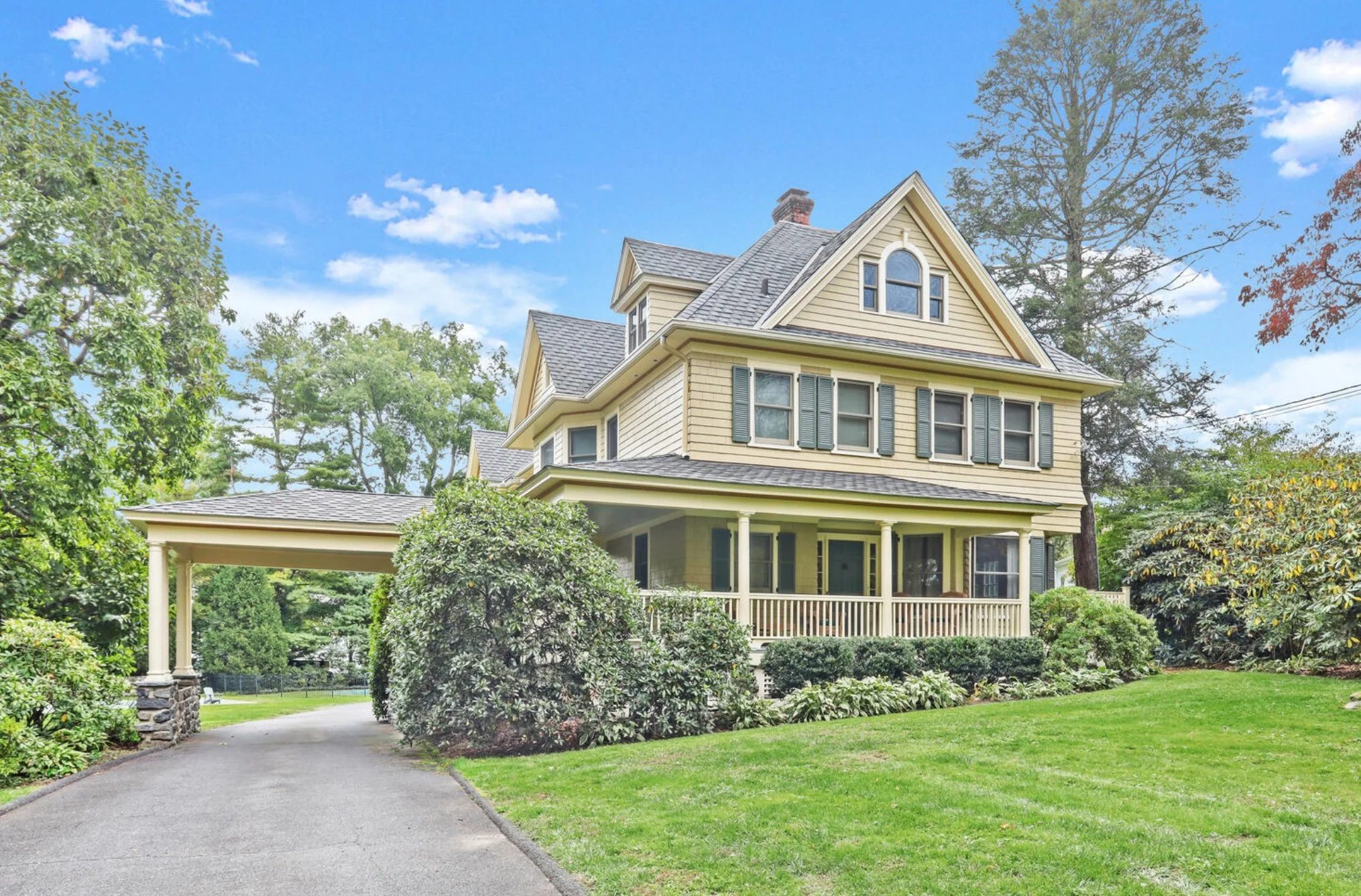 A large, yellow, three-story house with a front porch, surrounded by green trees and bushes, under a blue sky with some clouds.