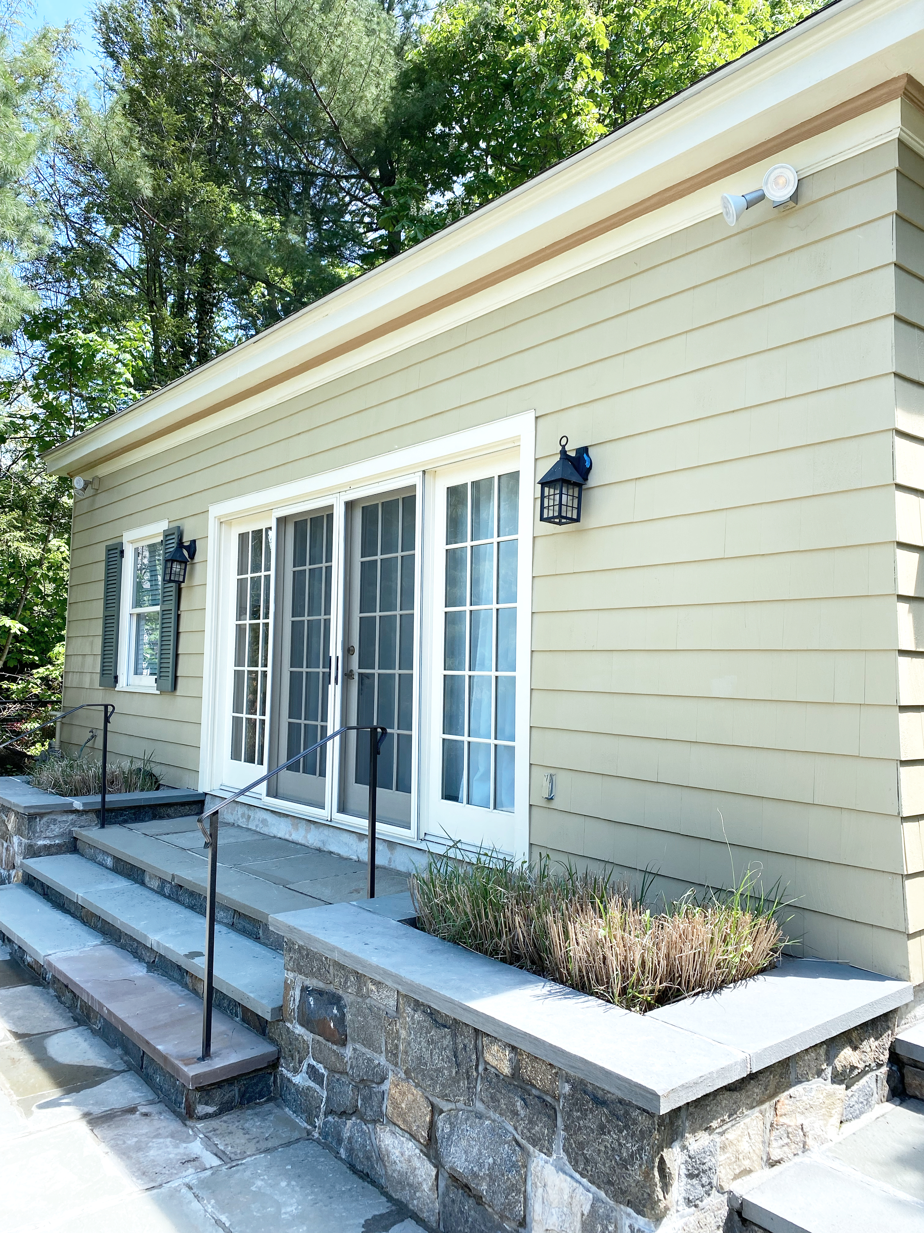 Yellow house exterior with stone steps leading to glass double doors, black handrails, window shutters, outdoor lighting fixtures, and planted flower beds, surrounded by green trees.