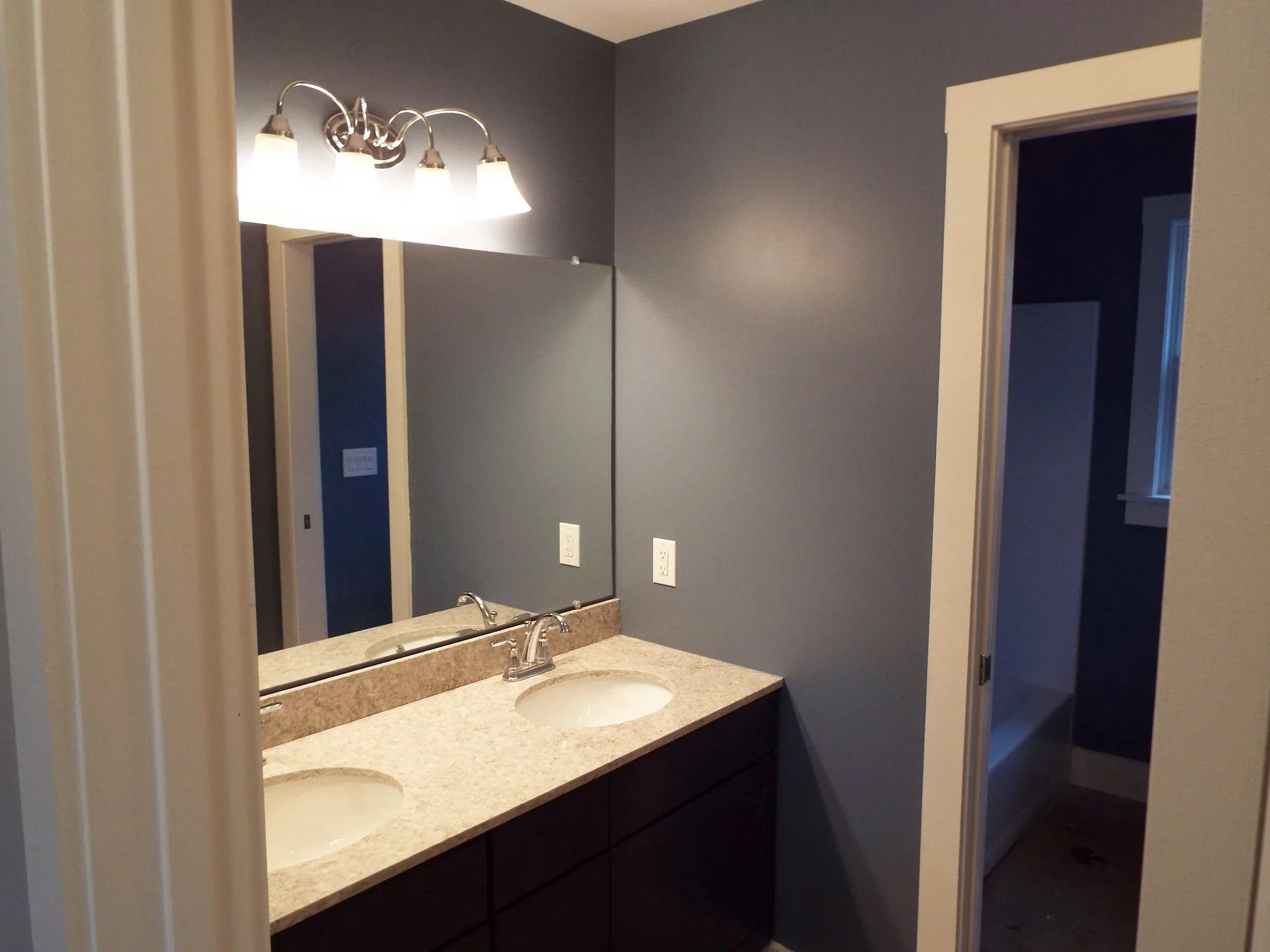 Bathroom with dual sink vanity, granite countertop, large mirror, blue walls, and a light fixture with four bulbs.
