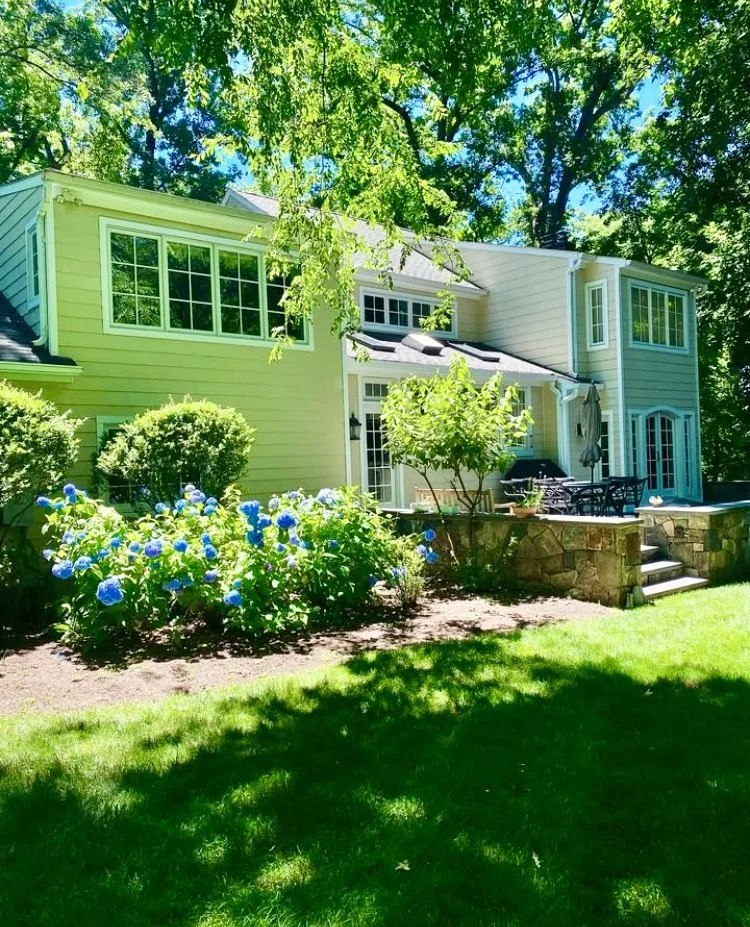 A yellow two-story house with large windows, surrounded by green trees, shrubs, and a garden with blue hydrangeas in bloom, a small patio with outdoor furniture, and a grassy lawn in the foreground.