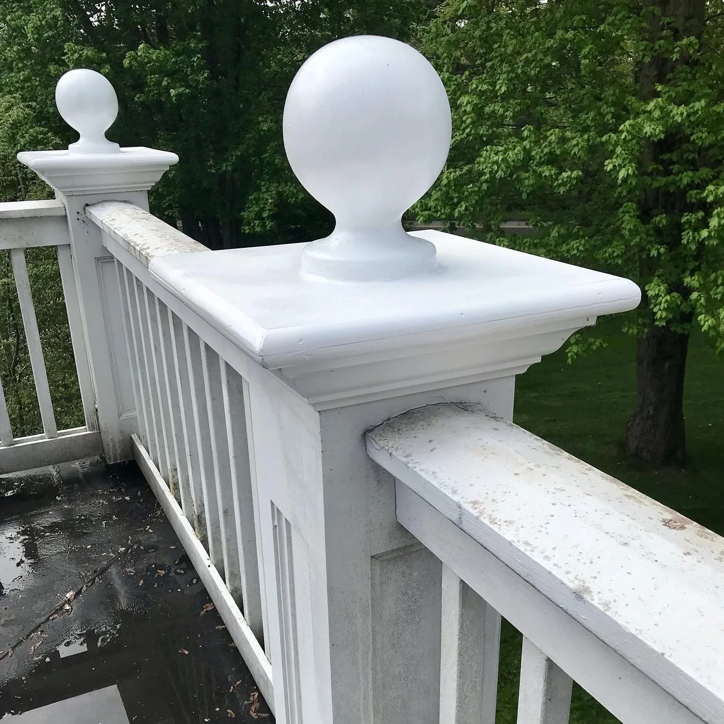 Close-up of a white wooden railing with decorative posts topped with spherical finials, showing some weathering and dirt, set against a background of green trees and grass.