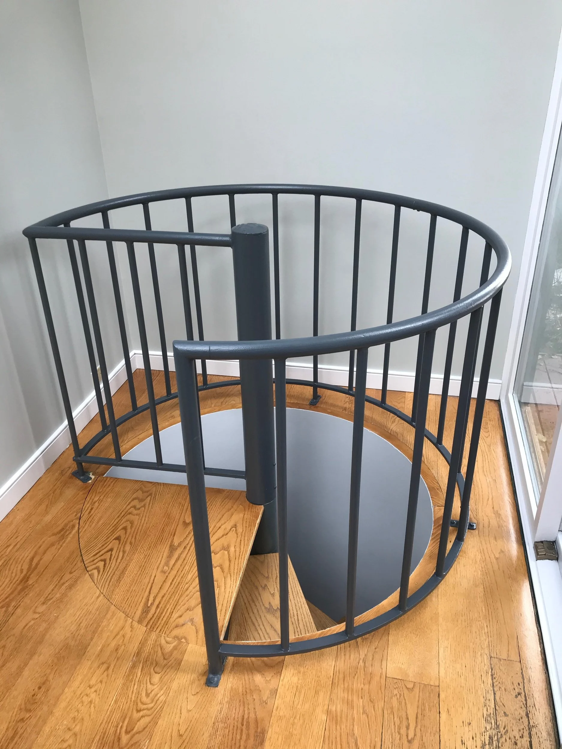 Spiral staircase with wooden steps and gray metal railing, inside a corner near a large window.