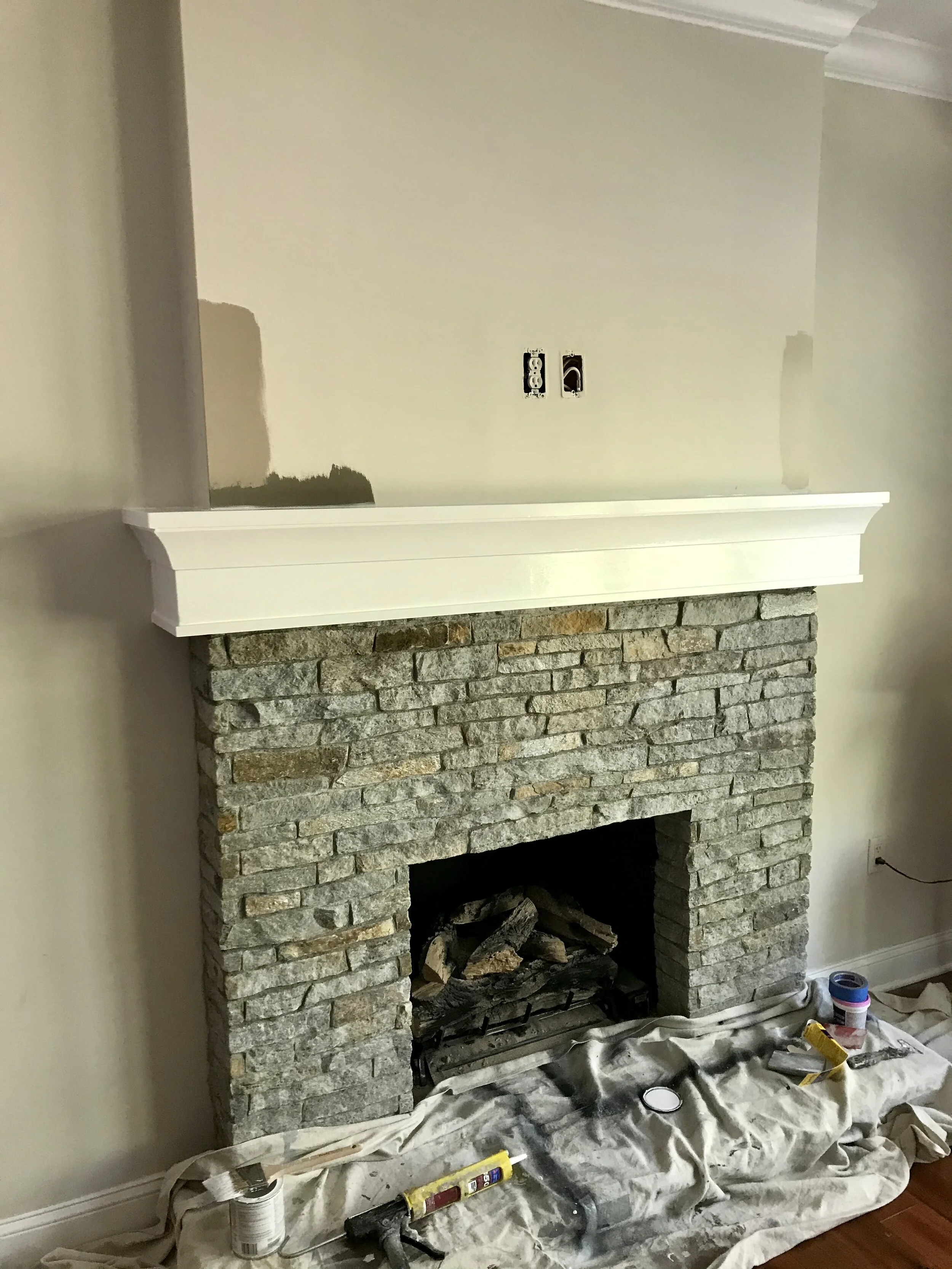 A stone fireplace with a white wooden mantel in the process of being painted, with painting supplies and painter's cloth spread on the floor in front.