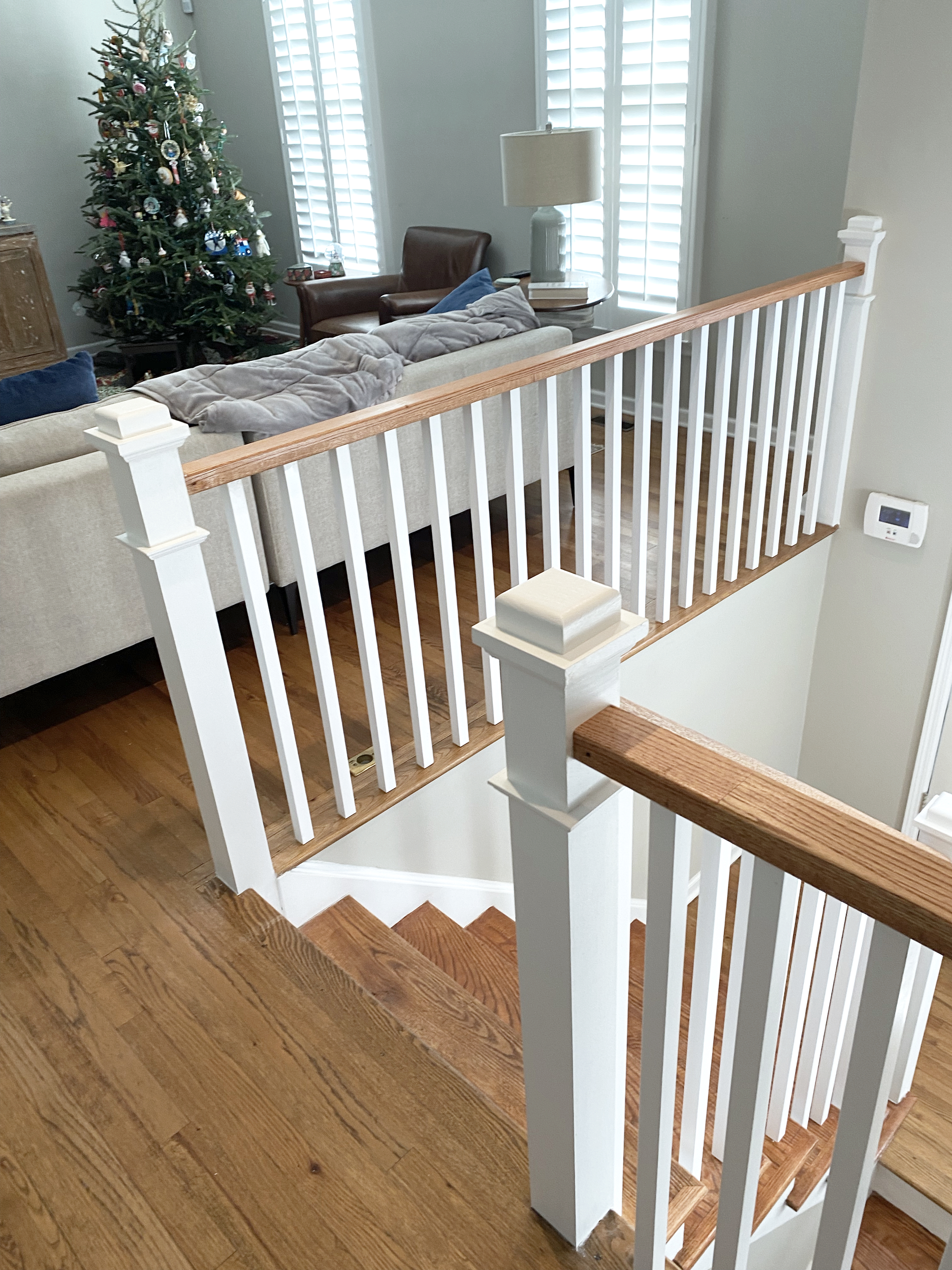 View of a staircase with a wood handrail and white balusters overlooking a living room with a Christmas tree, a beige sofa, a brown armchair, a floor lamp, and window shutters.