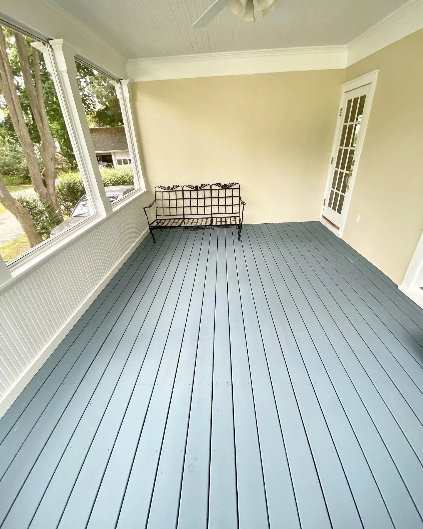 A porch or sunroom with blue painted wooden floorboards, cream-colored walls, large windows, a black metal bench, and a white ceiling fan.