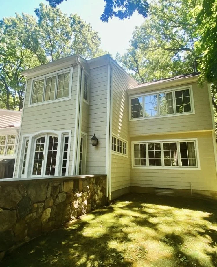 View of a house with cream siding, multiple large windows, and a stone wall in a wooded area on a sunny day.