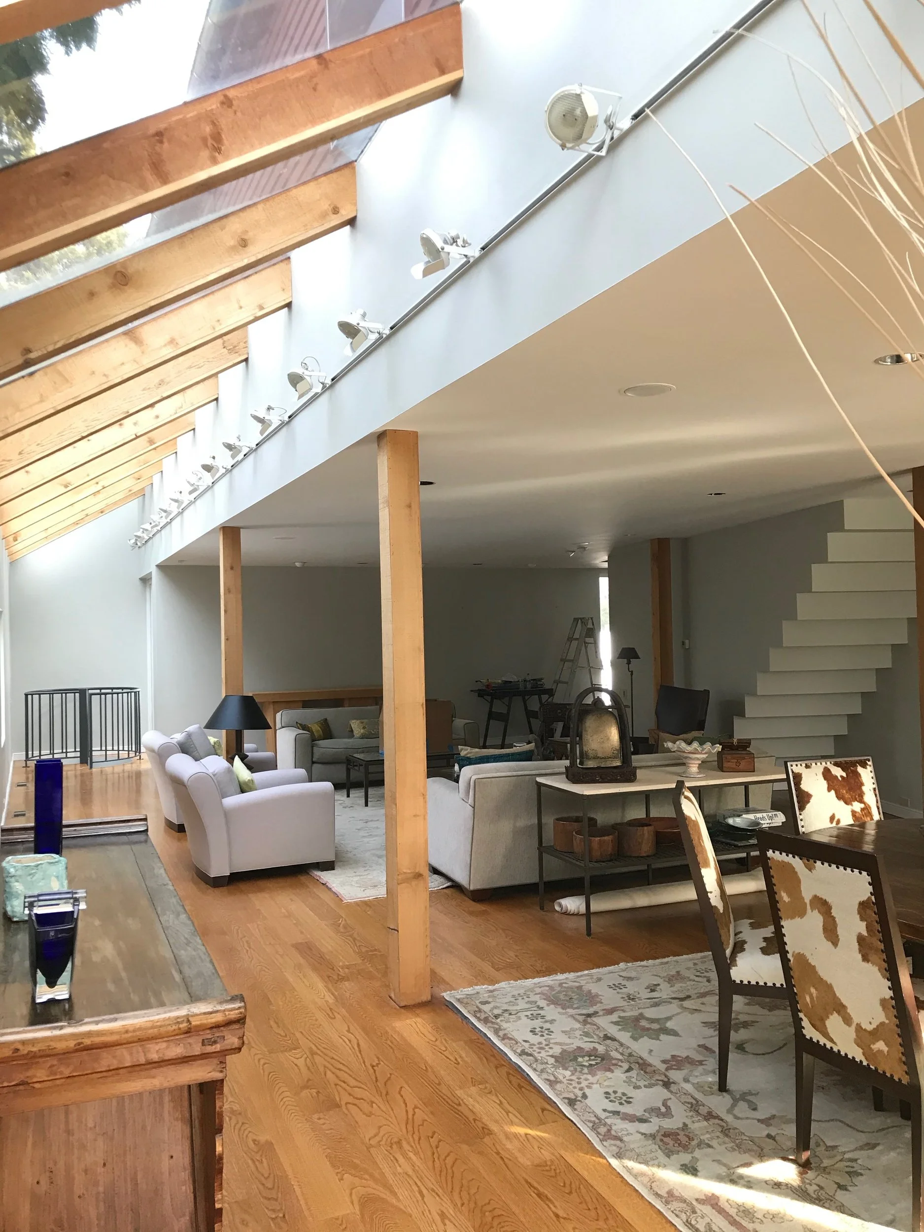 Open living room with white sofas, black lamps, and a wooden dining table with beige and brown patterned chairs. There are wooden beams on the slanted ceiling and a staircase on the right side. The room has hardwood floors and neutral-colored walls.