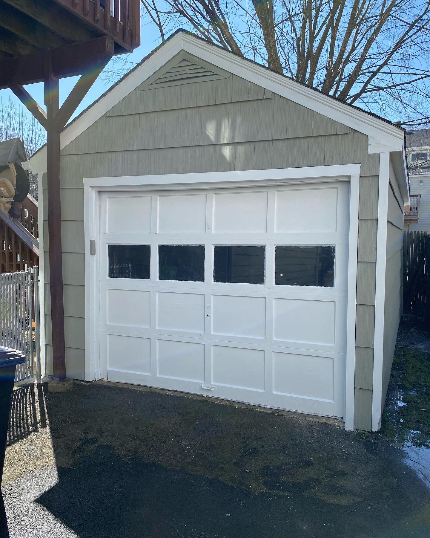 A small beige garage with a white sectional garage door that has four windows at the top. The garage is outdoors, with a tree behind it, a fence on one side, and a clear blue sky.