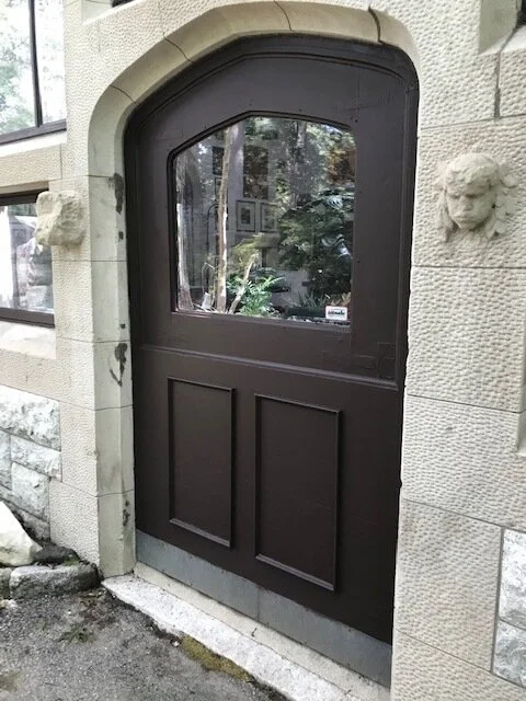 Photo of a brown wooden gate with glass window, built into a stone wall with lion head sculptures on both sides.
