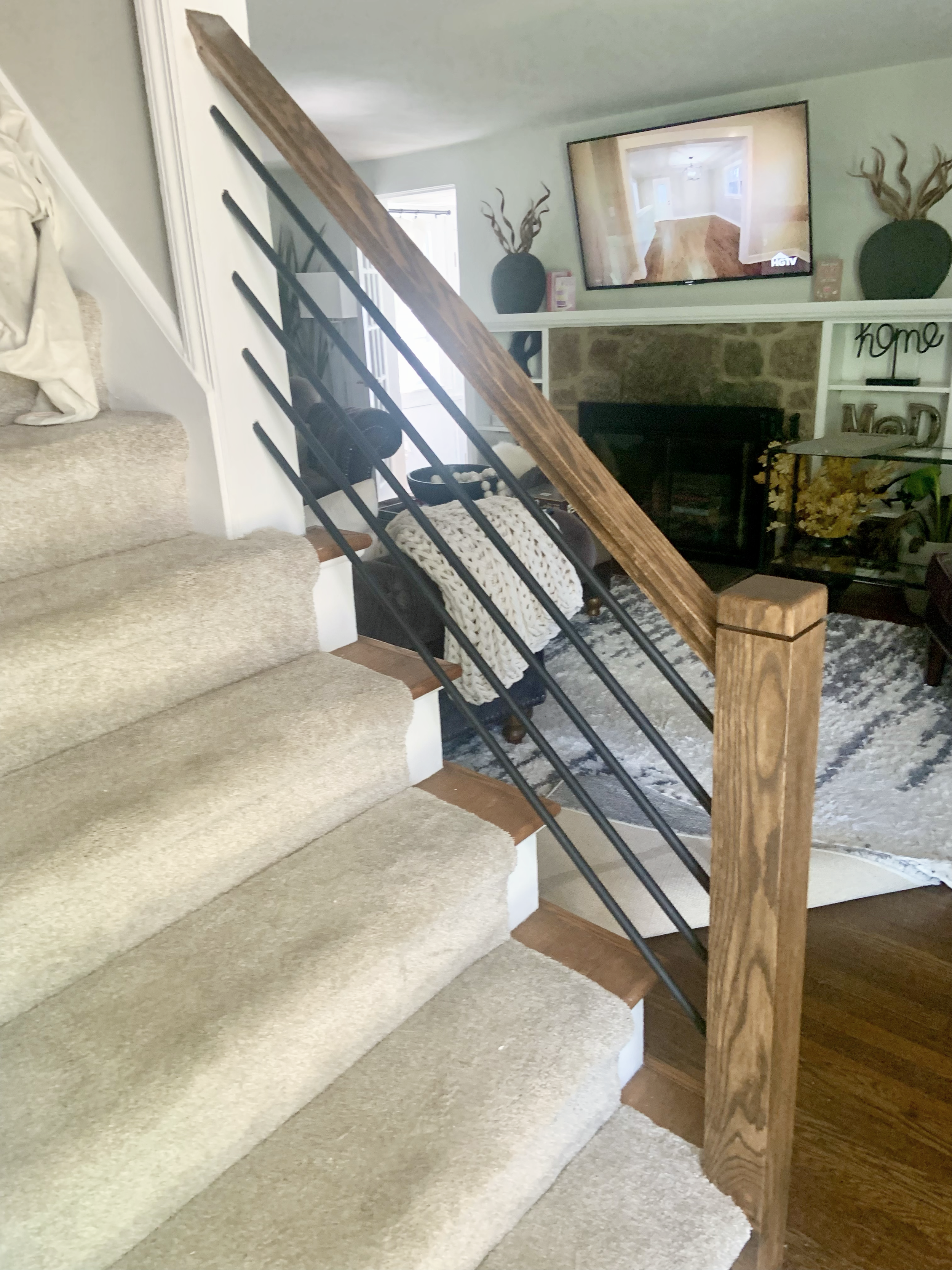 A staircase with beige carpeted steps and a wooden handrail with black metal balusters in a living room. In the background, there is a wall-mounted TV, a fireplace, and decorative items.