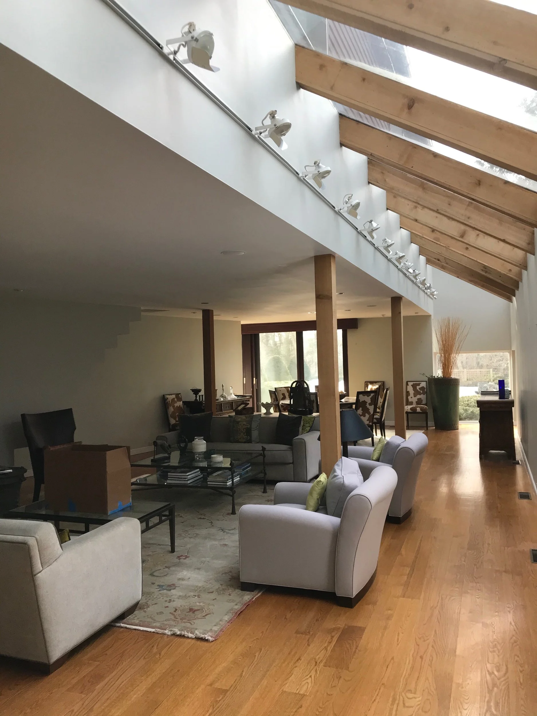 Living room with wooden floors, white and gray sofas, armchairs with green pillows, a glass coffee table with books, and a large window with sliding doors in the background. There is a sloped ceiling with skylights and a track lighting system.