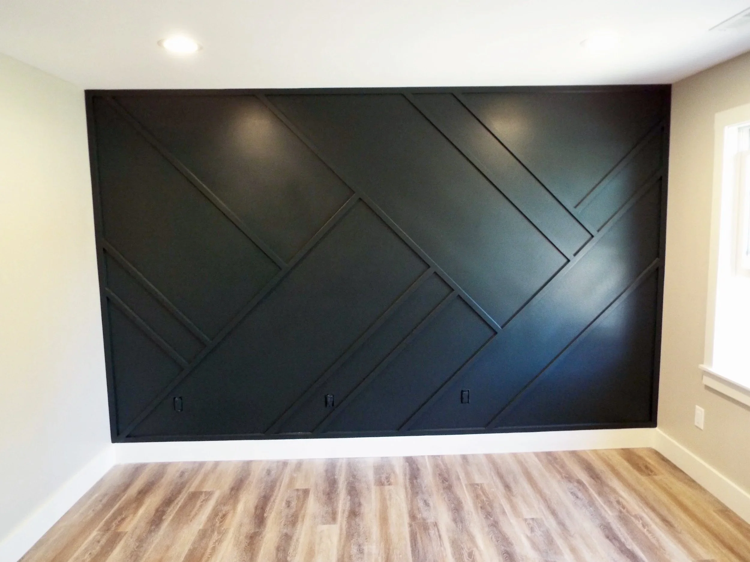 Empty room with a large black textured wall panel, light-colored walls, wooden flooring, and a window on the right side.