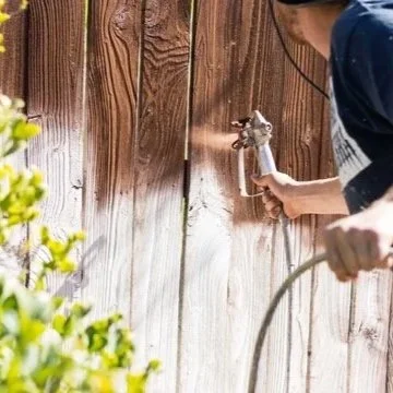 A person using a garden hose to spray water on a wooden fence.