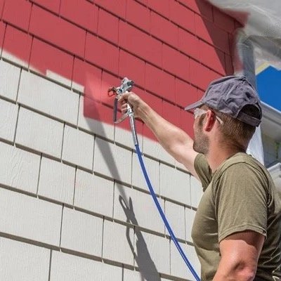 A man wearing a cap and sunglasses pressure washing the exterior wall of a house where the siding is being cleaned.