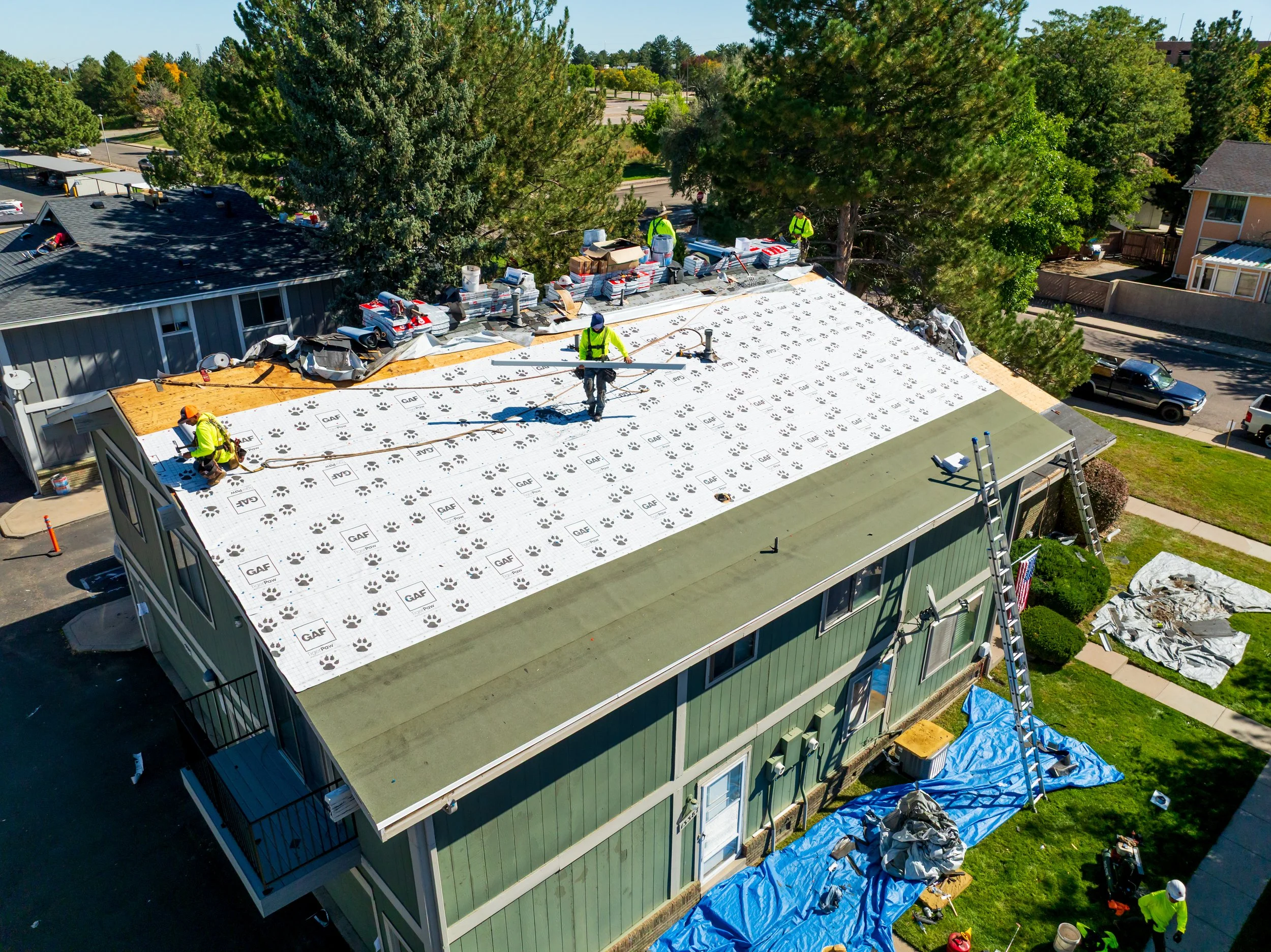 Construction workers installing a new roof on a green residential building, with roofing materials and equipment nearby, during daytime.