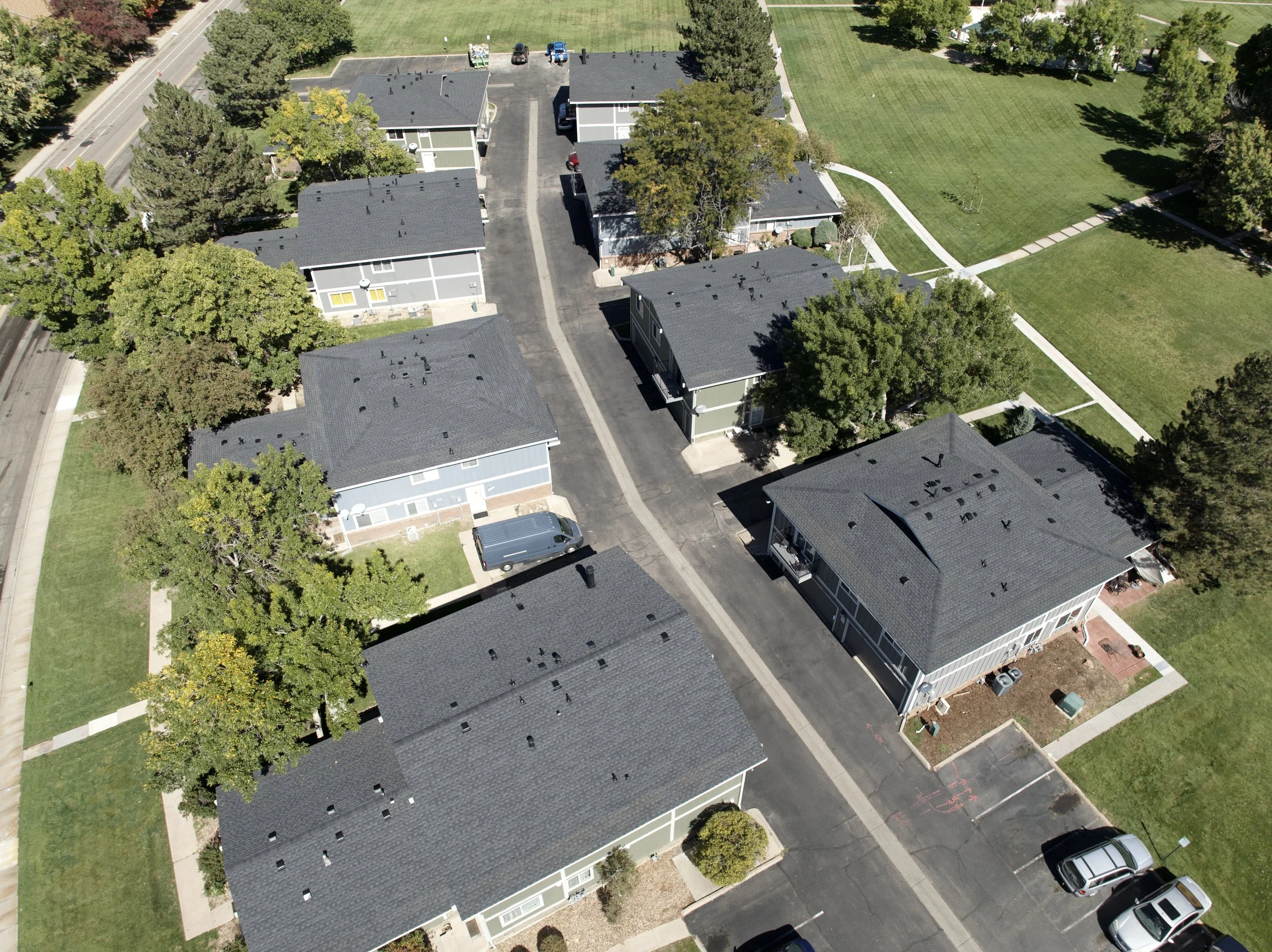 Aerial view of a residential apartment complex with multiple two-story buildings, parking spaces, and a large grassy park area with paved walking paths and trees.
