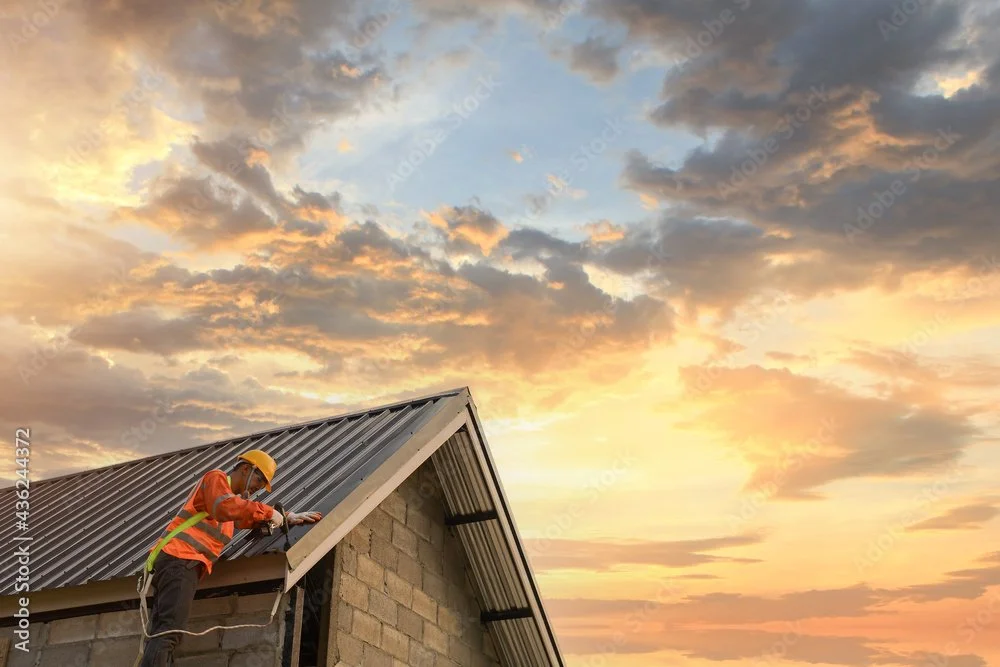 A construction worker wearing a yellow safety helmet, orange safety vest, and harness, working on the roof of a building during sunset.