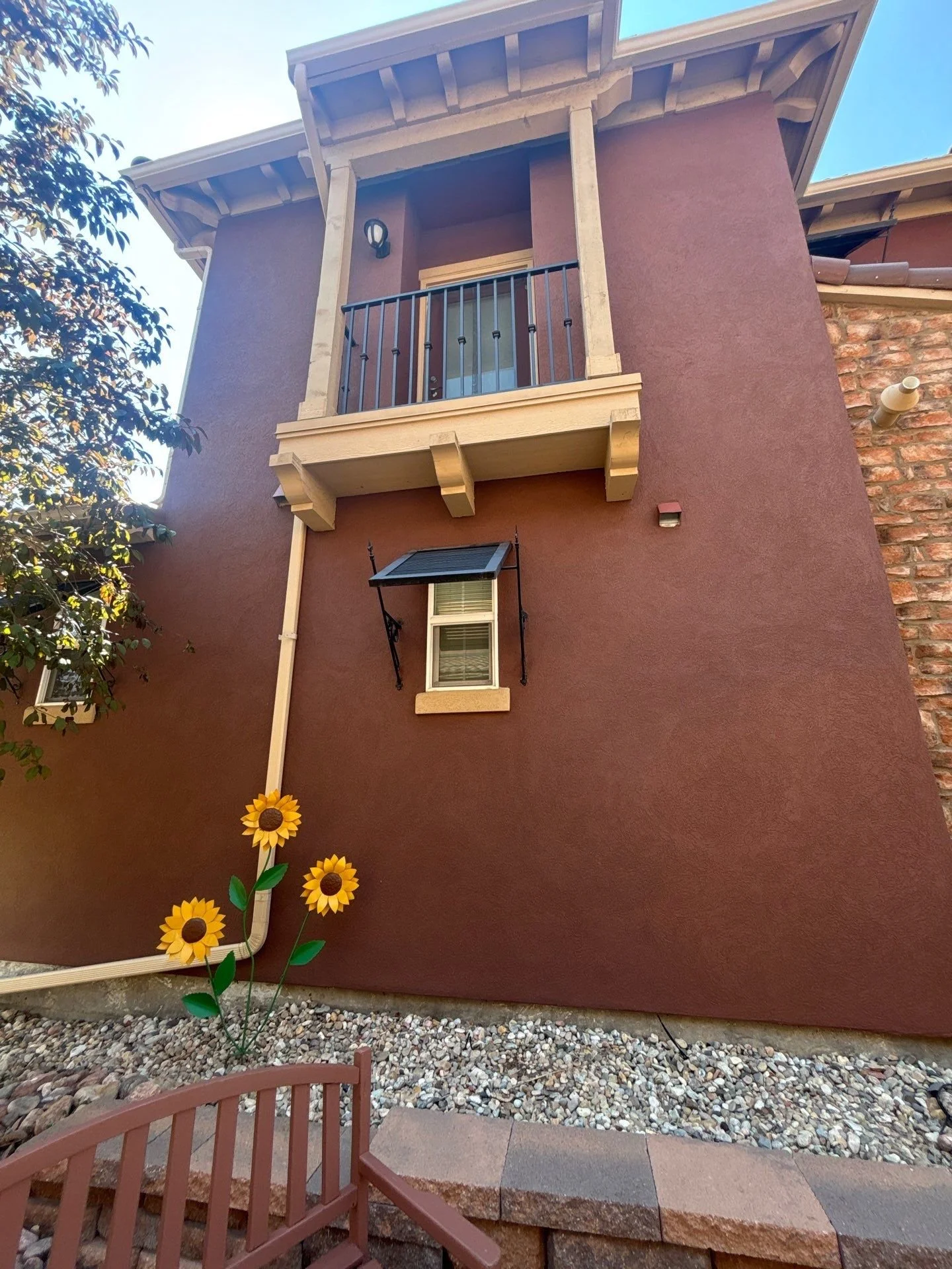 The image shows the exterior of a multi-story house with a reddish-brown stucco wall. There is a small balcony on the second floor with a black metal railing and a light fixture next to the balcony door. Below the balcony, there is a window with an open window awning. The ground features decorative gravel, a terracotta-colored garden chair, and three artificial sunflowers near the house.