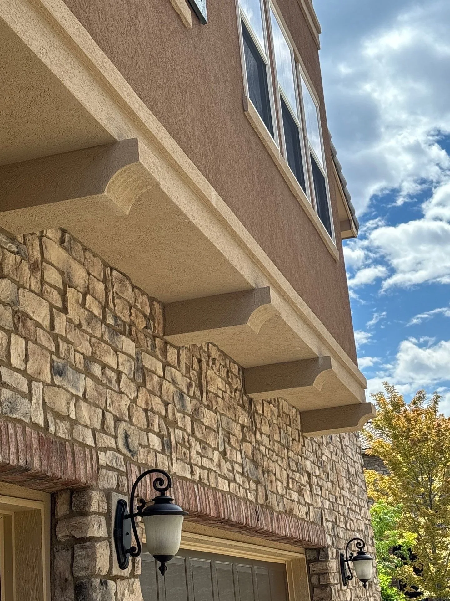 Close-up picture of the exterior of a multi-story house featuring a stone lower wall, beige trim, and several windows, with outdoor light fixtures and a partly cloudy sky.