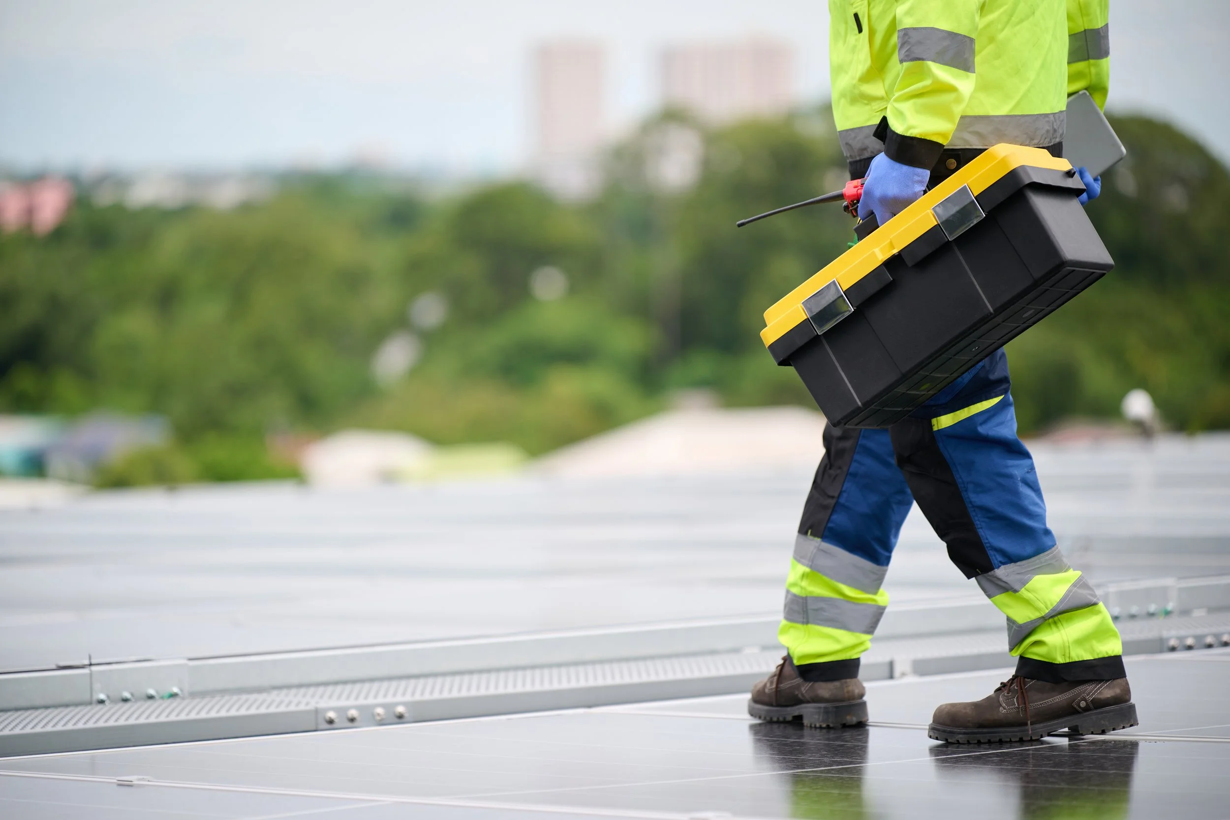 Worker inspecting solar panels on a rooftop, holding a toolbox and equipment.