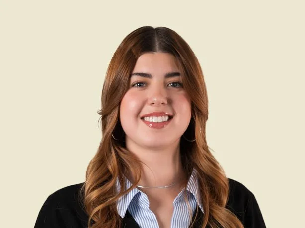 Portrait of a young woman with long, wavy brown hair, smiling, wearing a black blazer over a blue striped shirt, against a light background.
