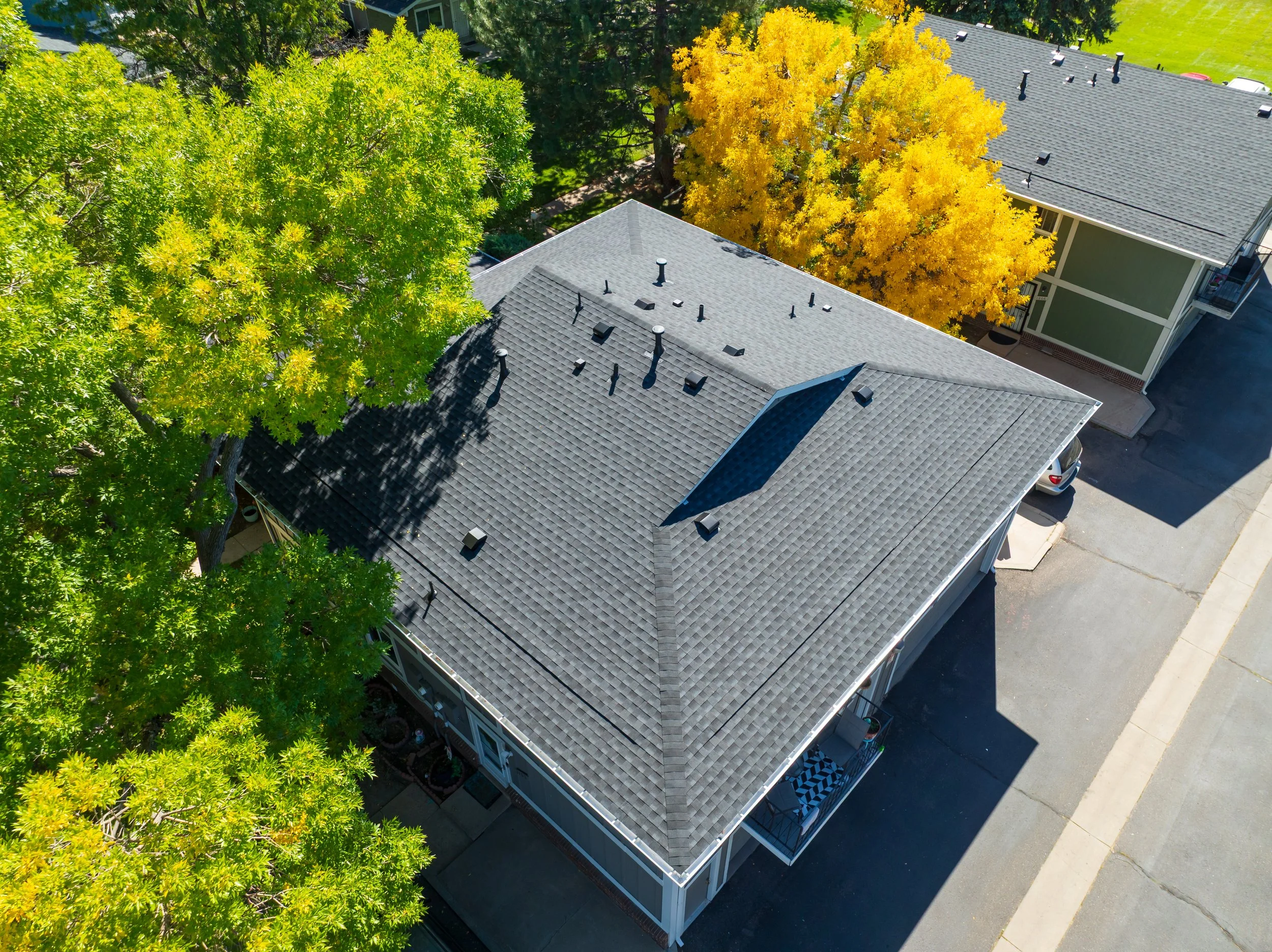 An aerial view of a house with a gray shingled roof, surrounded by green and yellow autumn trees, adjacent to a paved street and parking area.