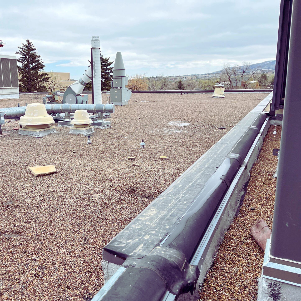 Rooftop with various ventilation and HVAC equipment, including vents, ducts, and exhaust pipes, with a cloudy sky and distant trees and mountains in the background.