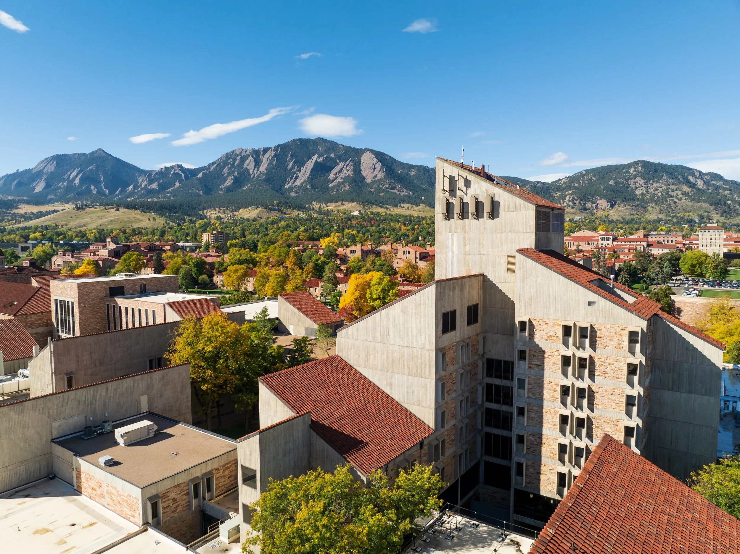 View of buildings with red tile roofs and a tall, modern concrete building, set against a background of mountains and a blue sky with a few clouds.
