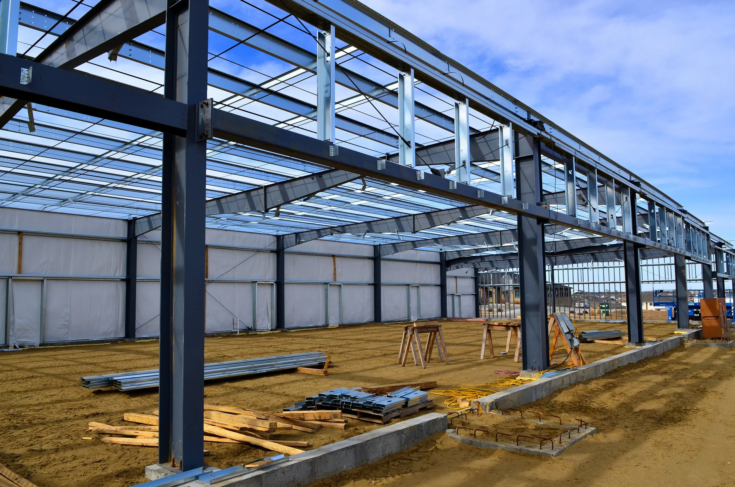 Steel framework of a building under construction on a dirt ground with construction tools and materials scattered around, under a partly cloudy sky.