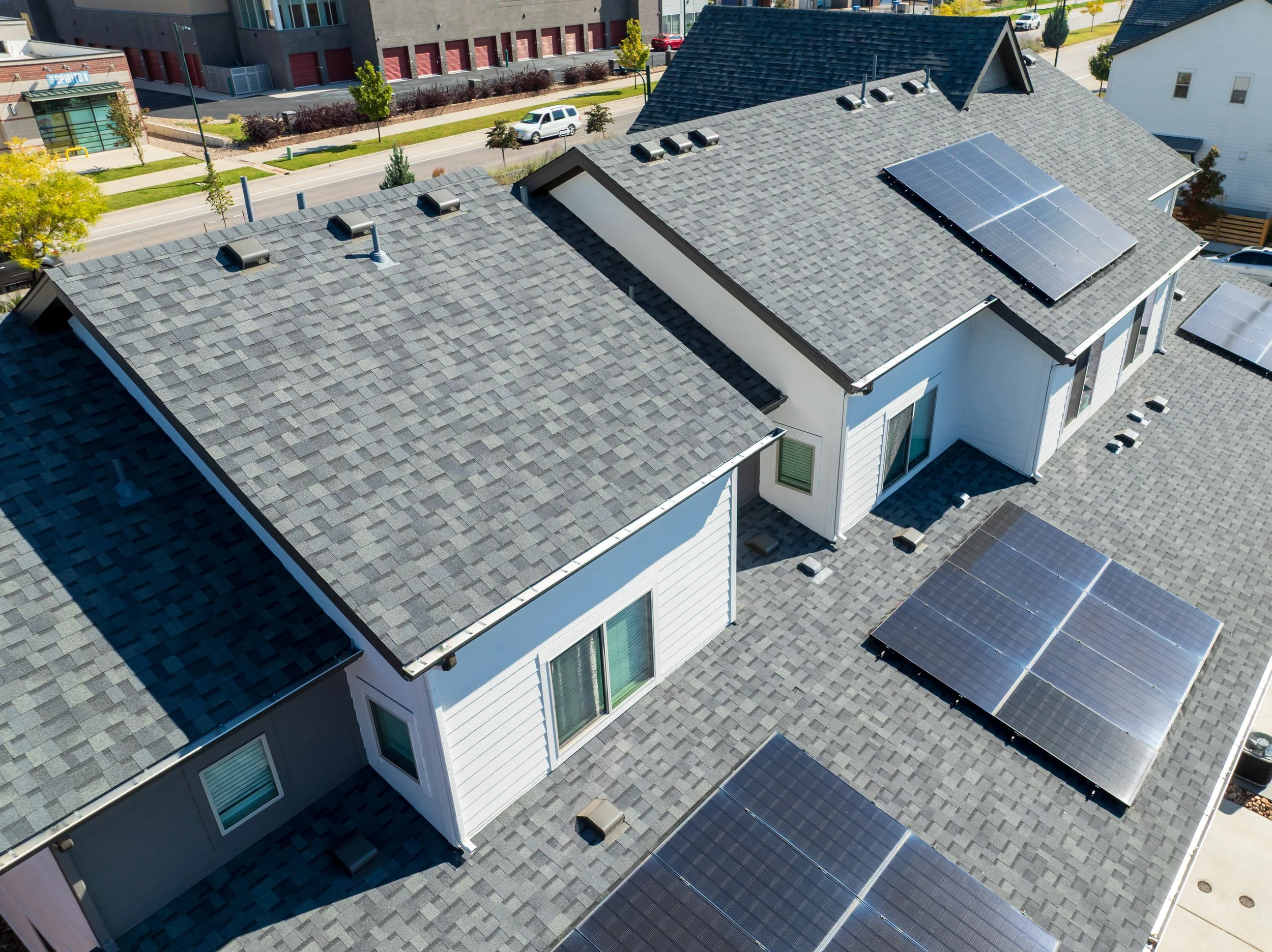 An aerial view of multiple residential rooftops with gray shingles, some featuring solar panels. A street with cars and trees is visible in the background.
