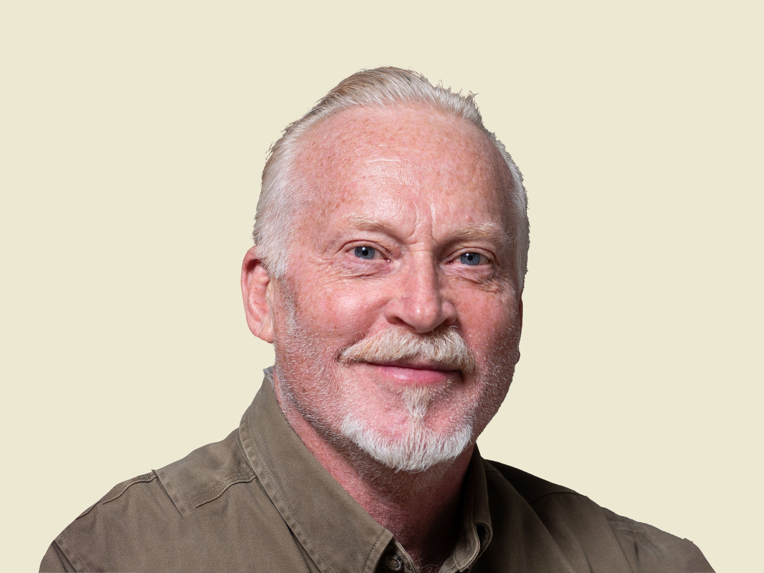 Close-up portrait of an older man with white hair, a beard, and mustache, wearing a brown shirt, smiling slightly, against a plain light background.