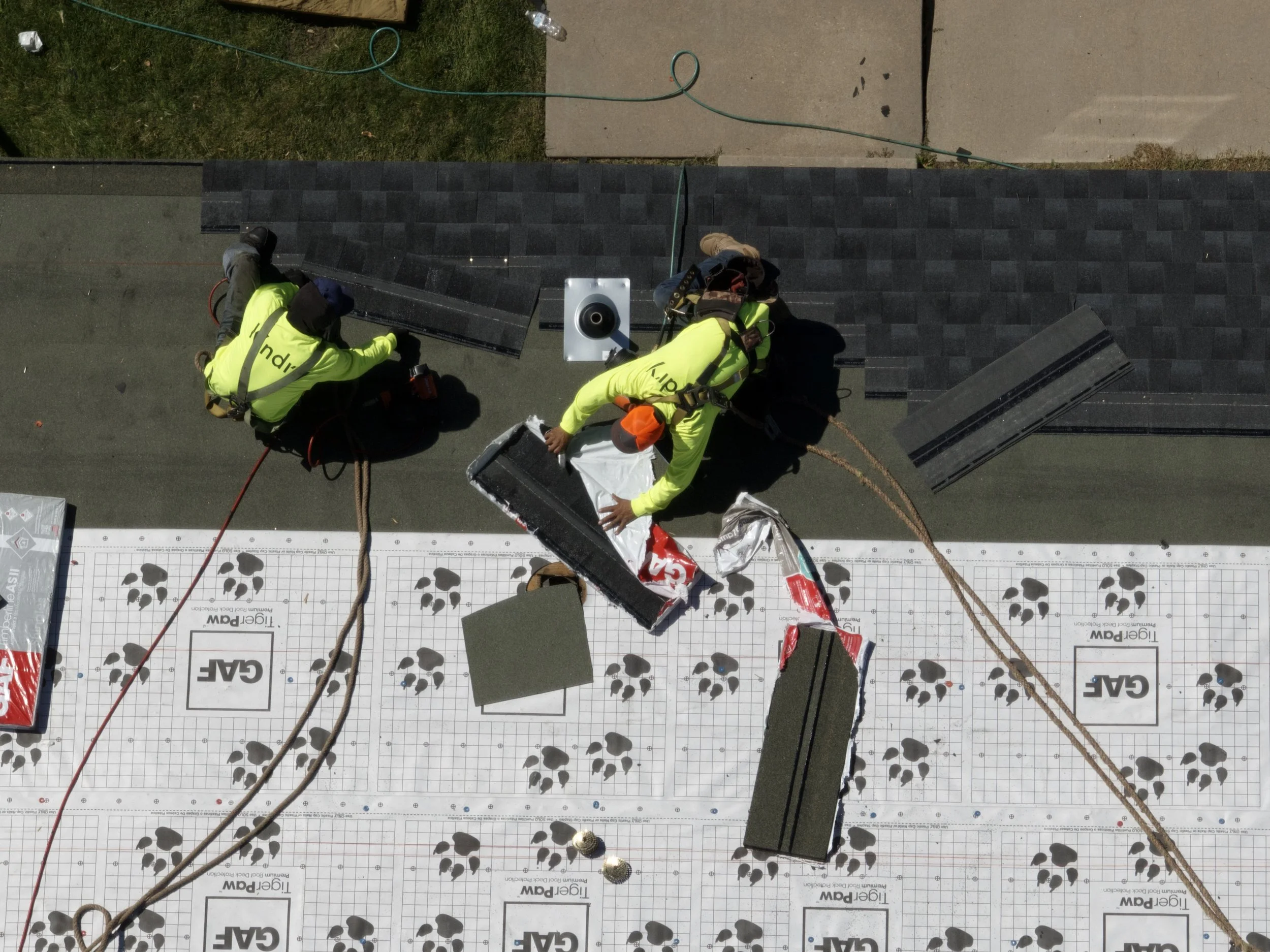 Two workers in yellow safety jackets working on roofing installation on a building with black shingles, using tools and materials. One worker is kneeling, the other is bent down arranging insulation or underlayment.