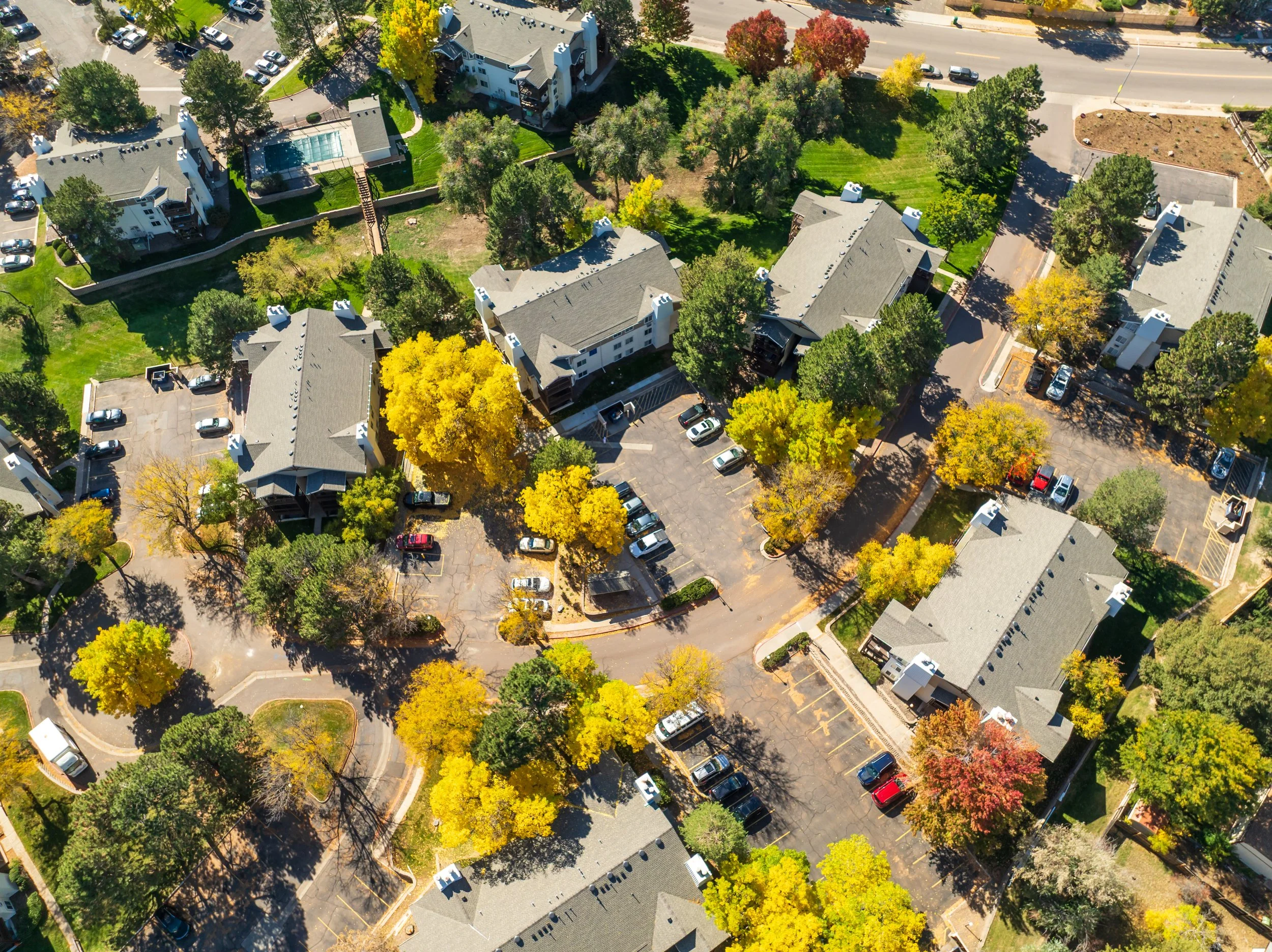 Aerial view of a residential apartment complex with multiple buildings, parking lots, and green trees with fall foliage.