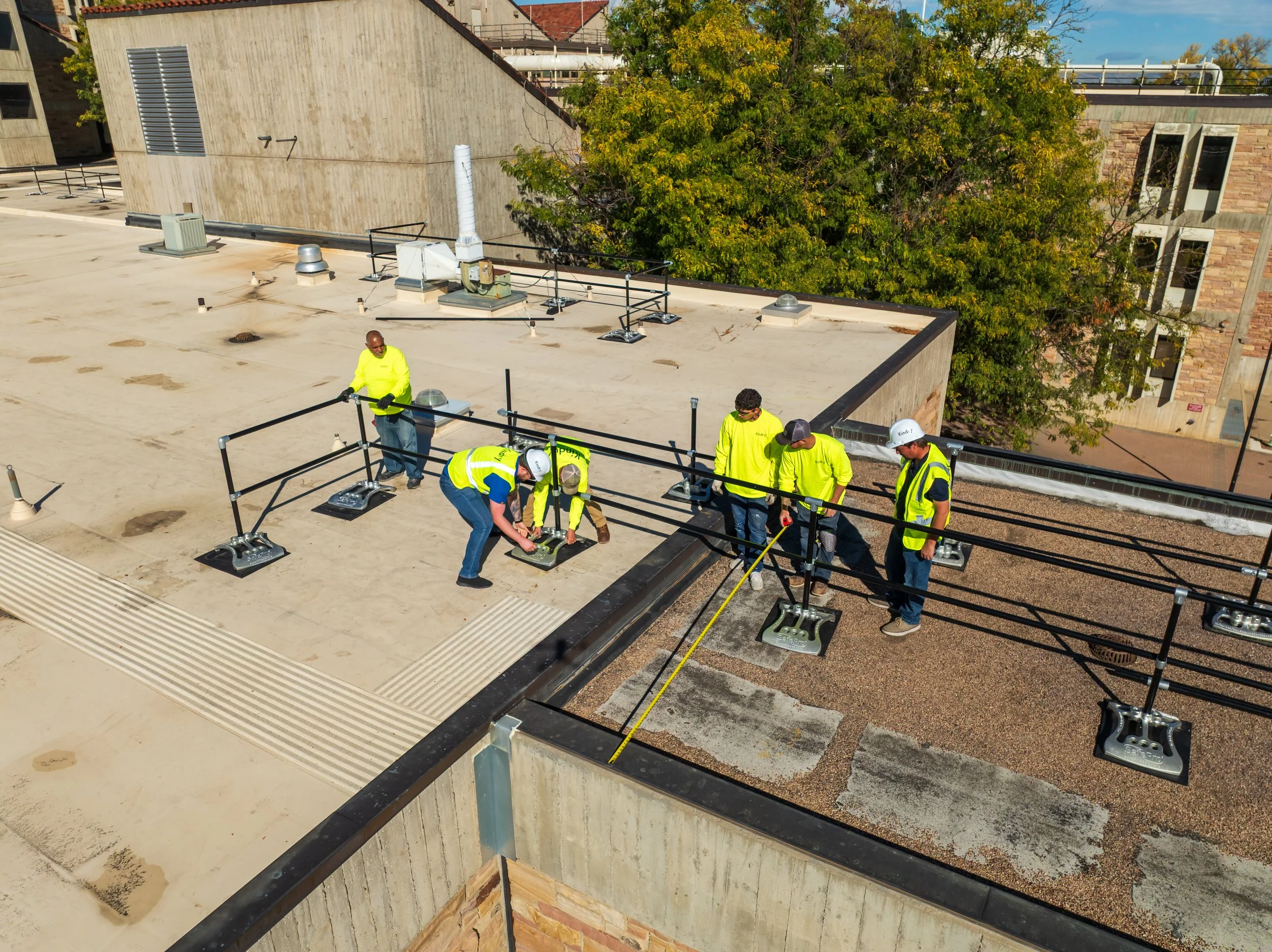 Group of construction workers wearing high-visibility safety vests and helmets working on the rooftop of a building. Some are handling equipment, and one appears to be measuring with a tape measure.