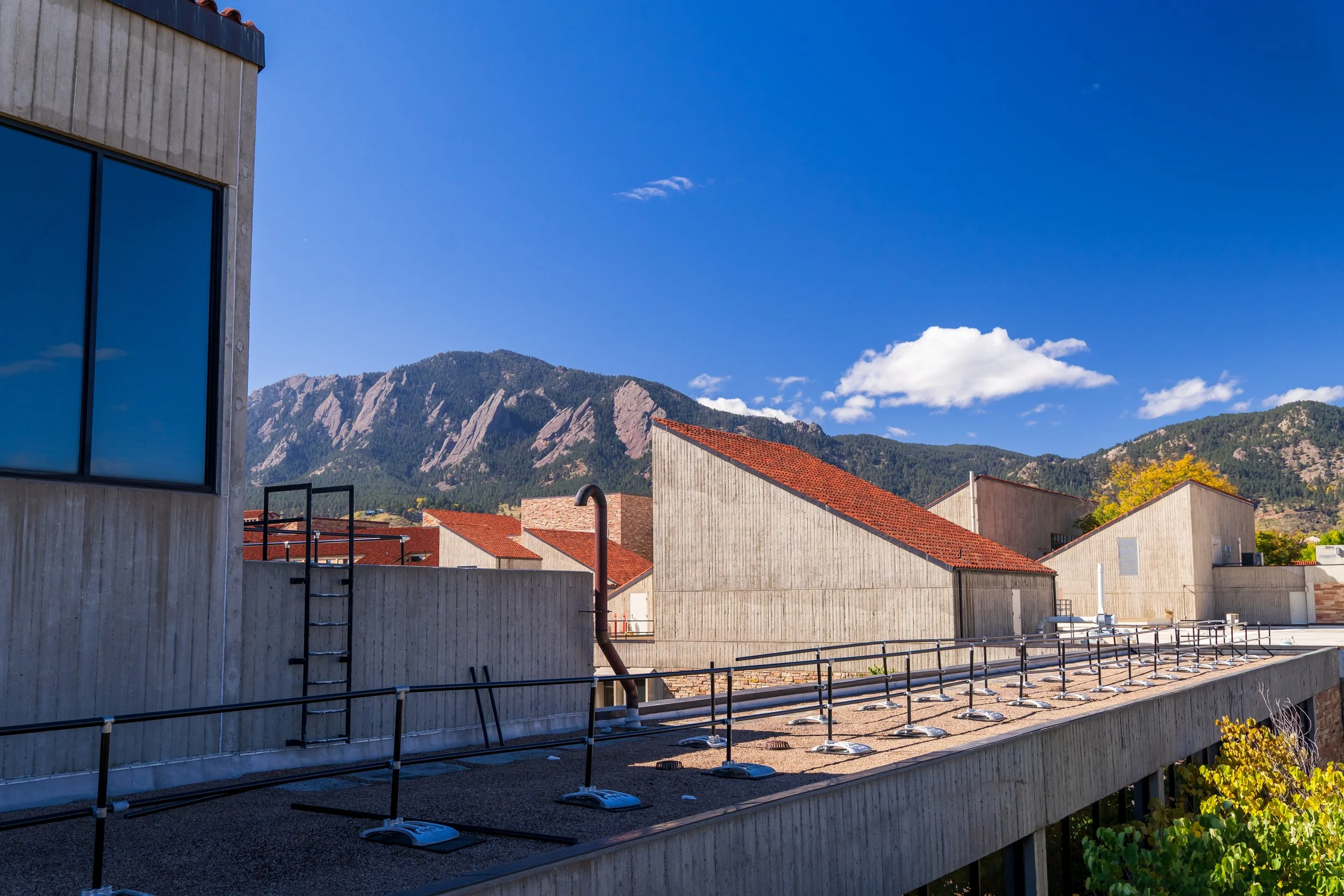 View of a modern building with concrete walls, red-tiled sloped roofs, and a railing on the rooftop, with mountains and a blue sky with clouds in the background.
