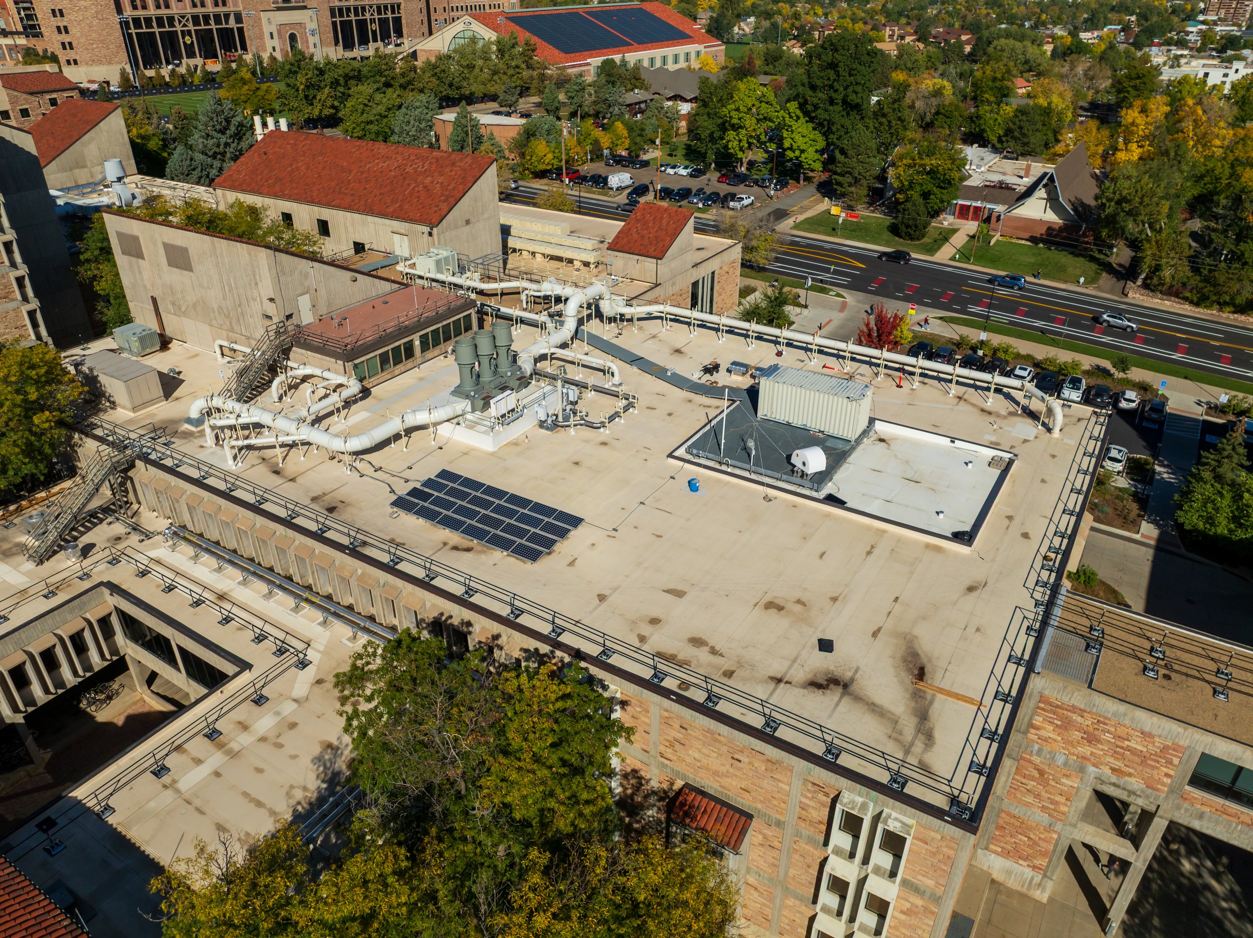 Aerial view of a building rooftop with HVAC equipment, pipes, solar panels, and a small rooftop structure, with streets and trees in the background.