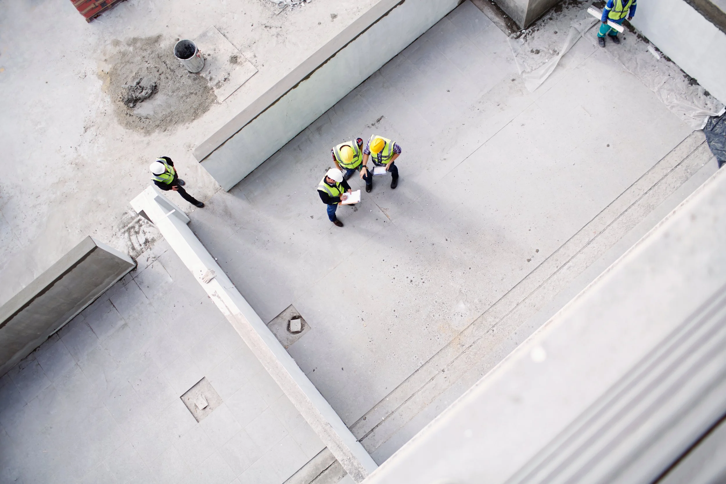 Top-down view of four construction workers wearing safety gear, including helmets and reflective vests, gathered and discussing plans on a construction site with concrete surfaces and walls.