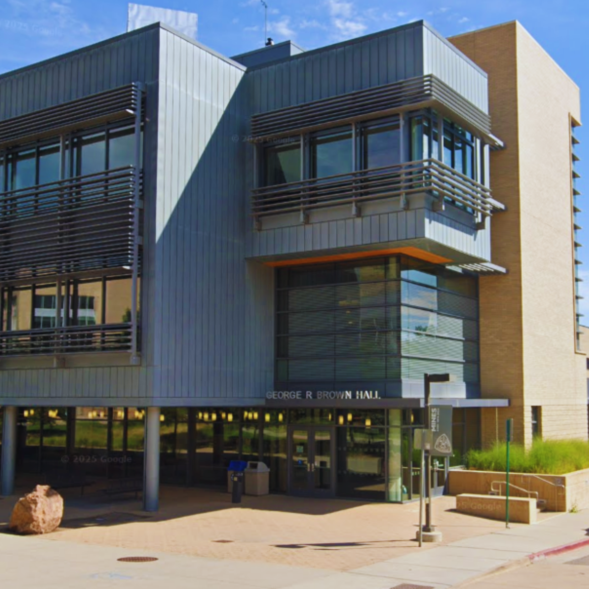Exterior of George R. Brown Hall, a modern building with metal panels, glass windows, and brown accents, with the entrance visible at street level under a canopy.