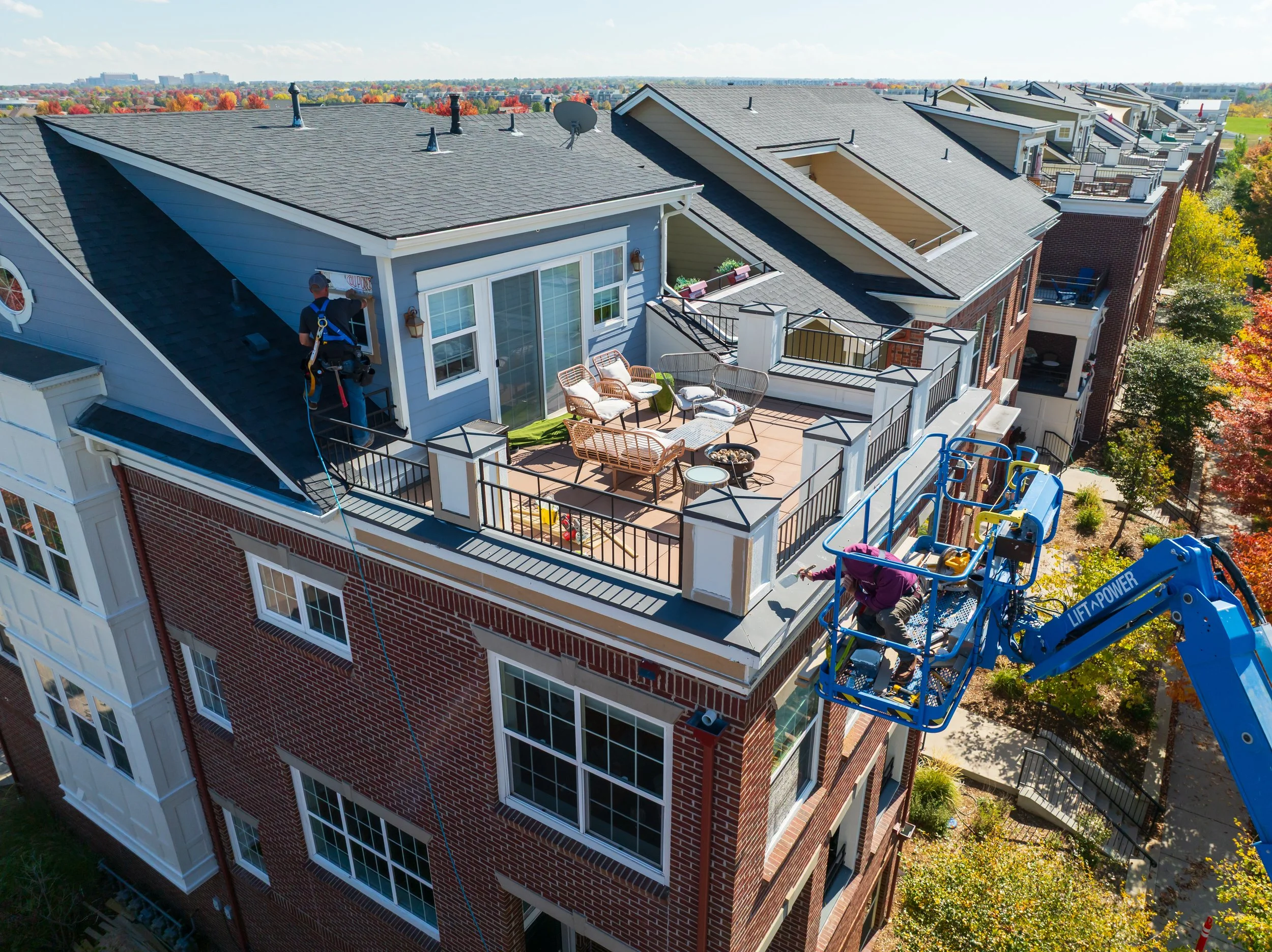 Workers cleaning the rooftop balcony of a multi-story residential building using a lift platform. The building has brick and siding exterior, large windows, and a patio with outdoor furniture. Trees with fall foliage are visible nearby and in the background.