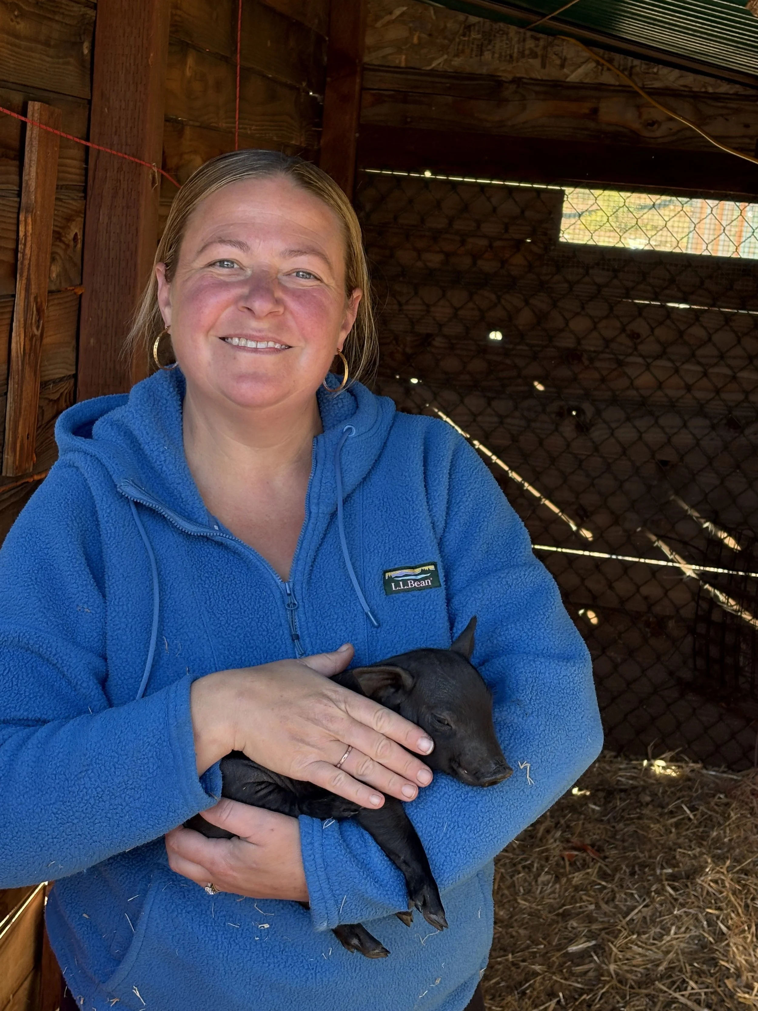 A woman smiling and holding a small black piglet inside a wooden barn, with sunlight filtering through a wire mesh window behind her.