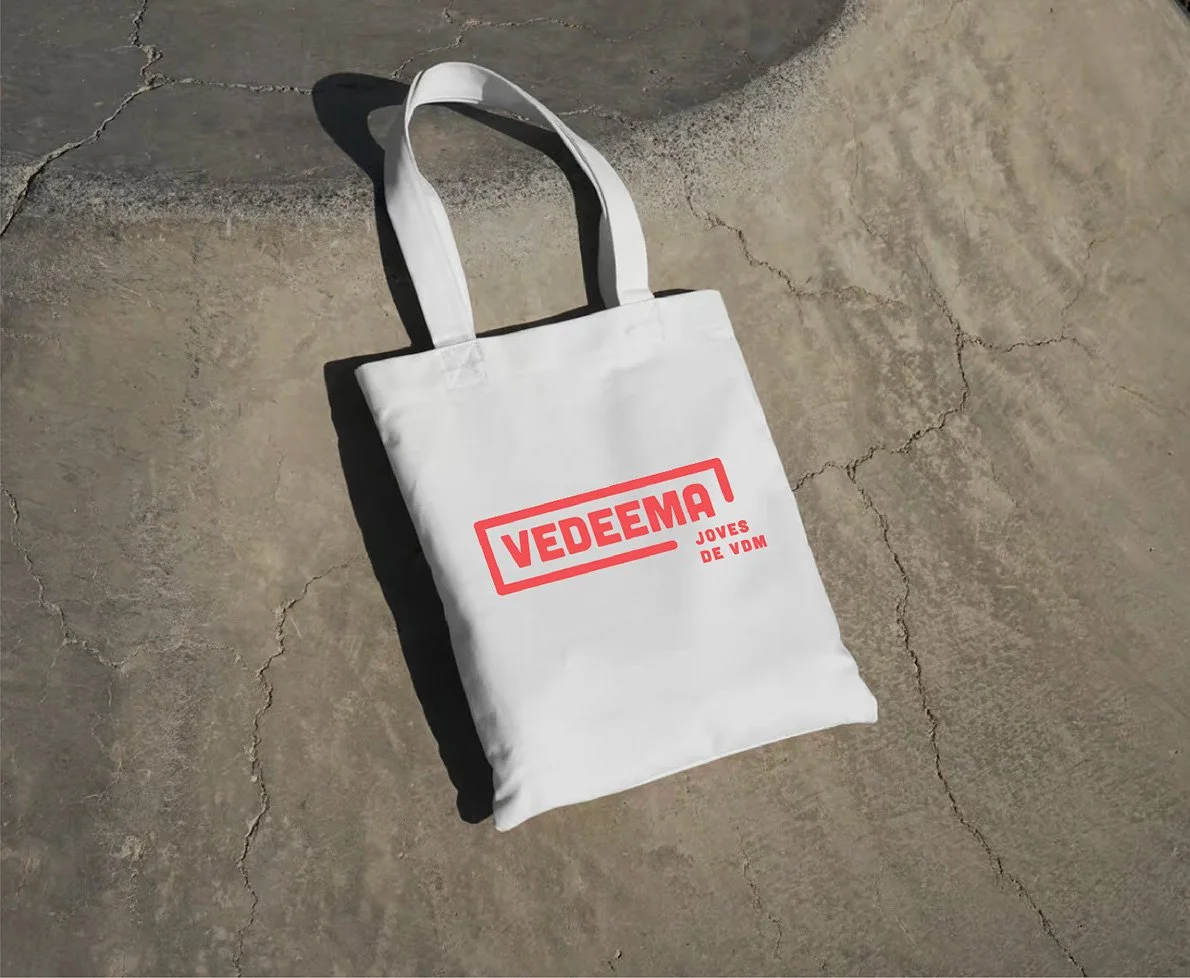 Una bolsa de tela blanca con las palabras en rojo 'VEDEMA JOVES DE VDM', colocada sobre una superficie de cemento con grietas y sombras_campaña identidad grafica de estudio berlin