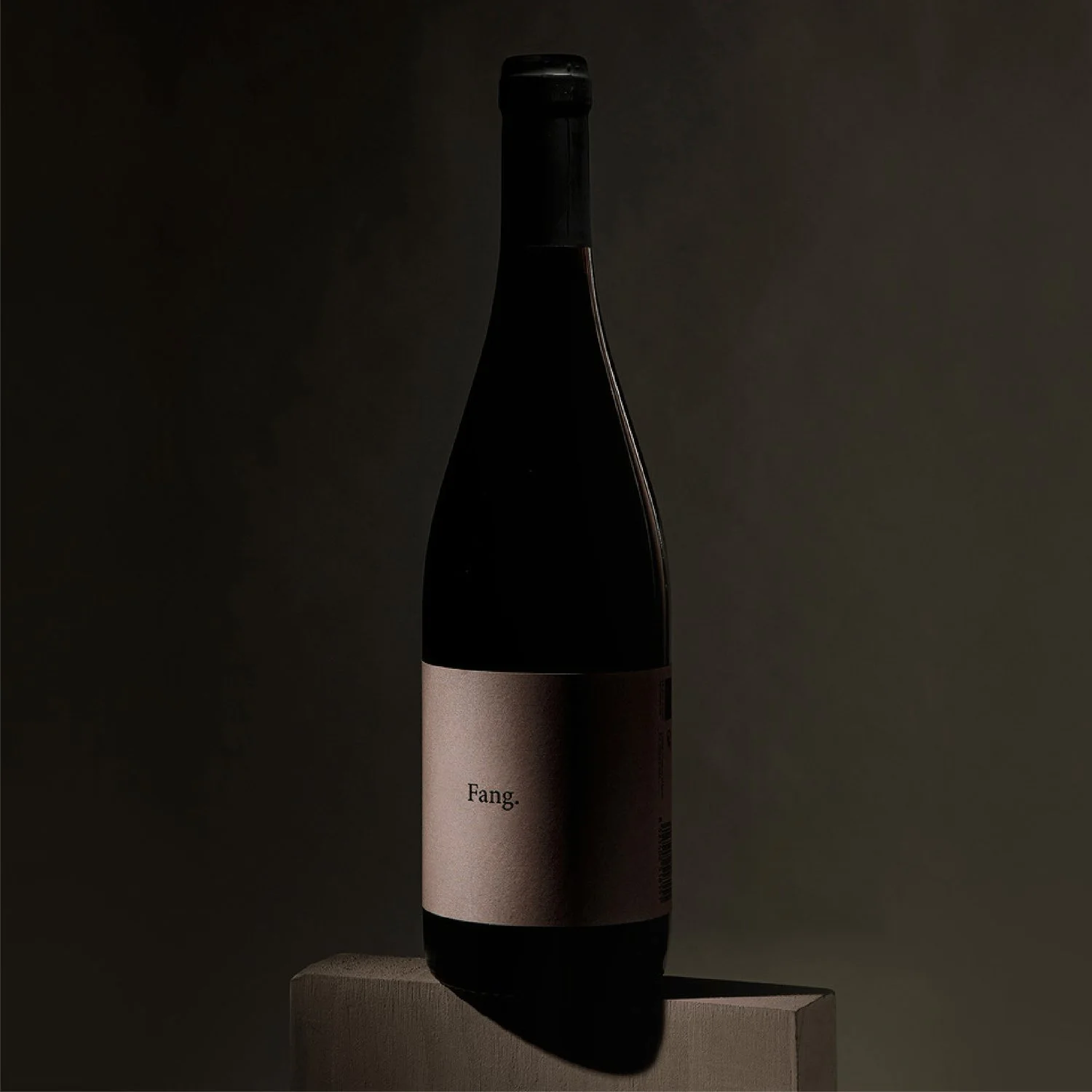Botella de vino negro con etiqueta beige que dice 'Fang', colocada sobre un soporte de madera contra un fondo oscuro_Viticultor del Penedès_Vi d’àmfora Fang_ diseño depackaging de botella vino echo por estudio berlin
