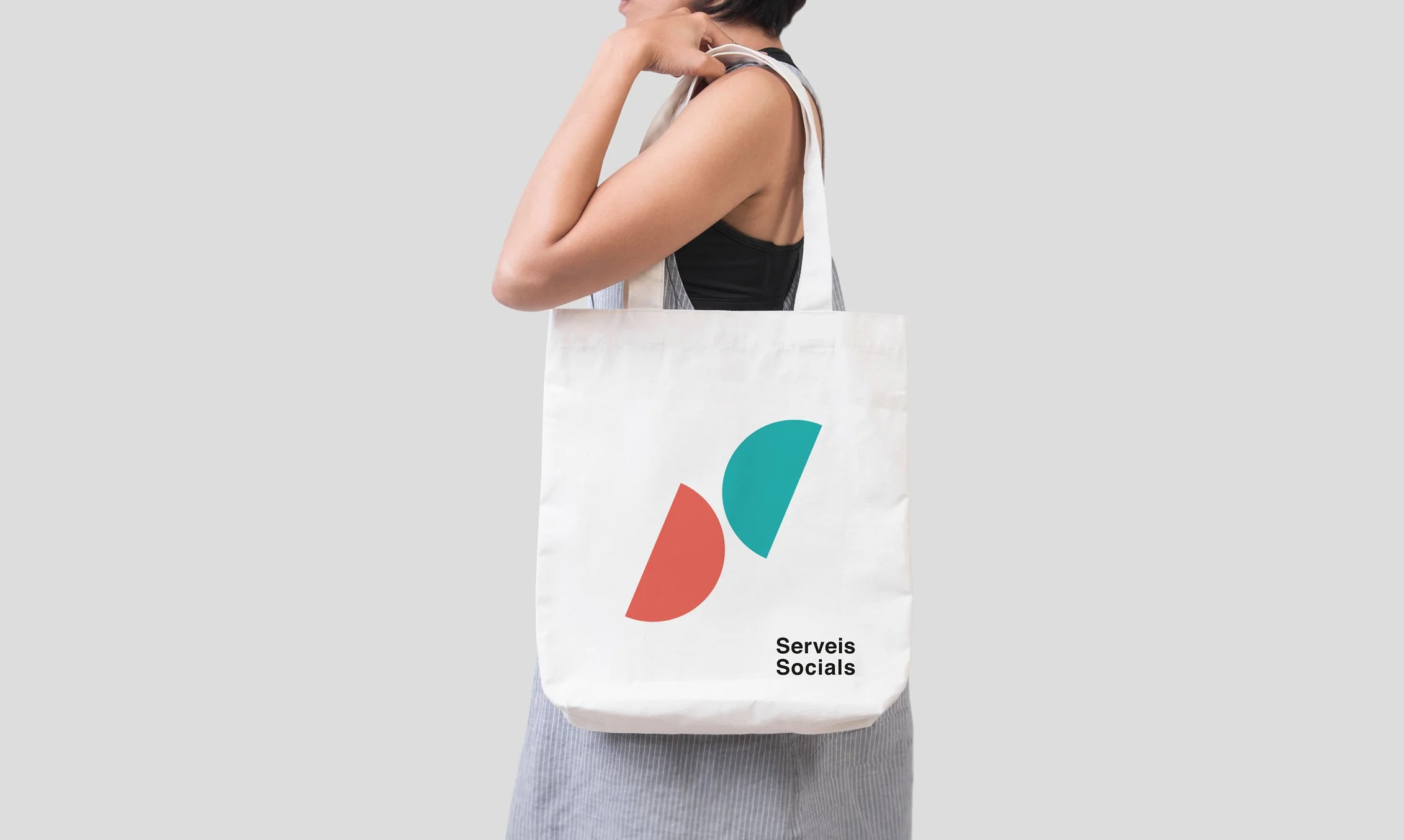 Mujer con camiseta negra y falda grisácea lleva una bolsa blanca con logotipo colorido y la leyenda 'Serveis Socials'_serveis socials_vilasar de mar_diseño identidad de estudio berlin