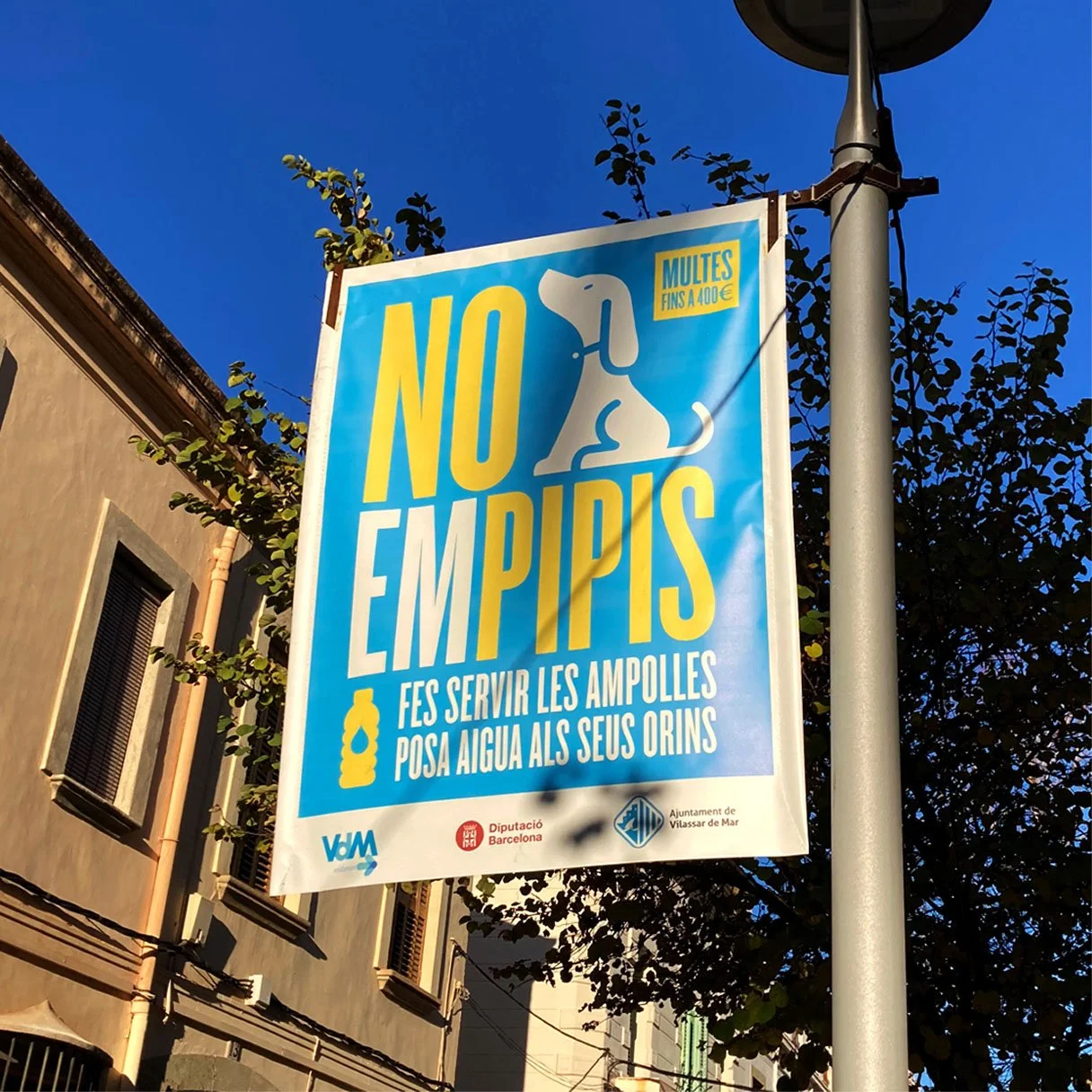 Cartel en la calle que dice "NO EMPIEPS" con dibujo de un perro y mensajes contra multas por no servir agua a las campanas de agua en Barcelona_campaña no empipis  per l'ajuntament de Vilassar de Mar_campaña identidad grafica de estudio berlin
