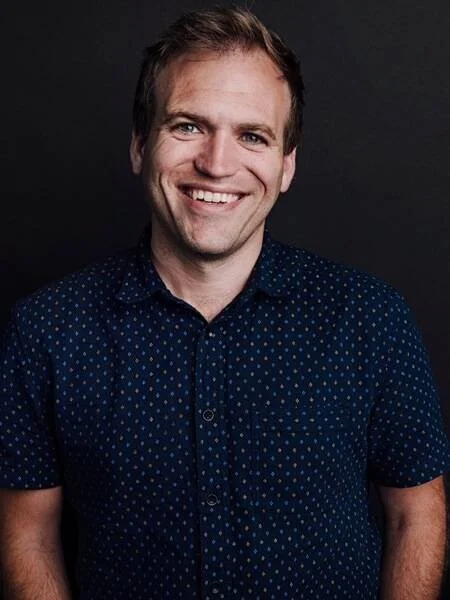 A smiling man with light brown hair, wearing a dark blue shirt with small white polka dots, against a dark background.