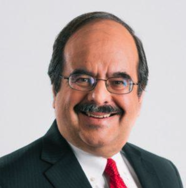 A man with glasses, a mustache, and dark hair, wearing a dark suit, white shirt, and red tie, smiling in front of a plain background.