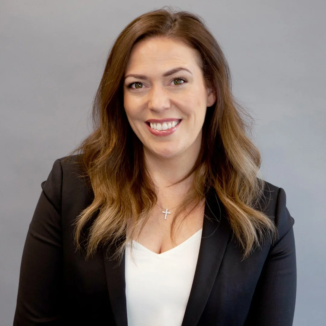 A smiling woman with shoulder-length brown hair wearing a black blazer and a white top, standing in front of a gray background.