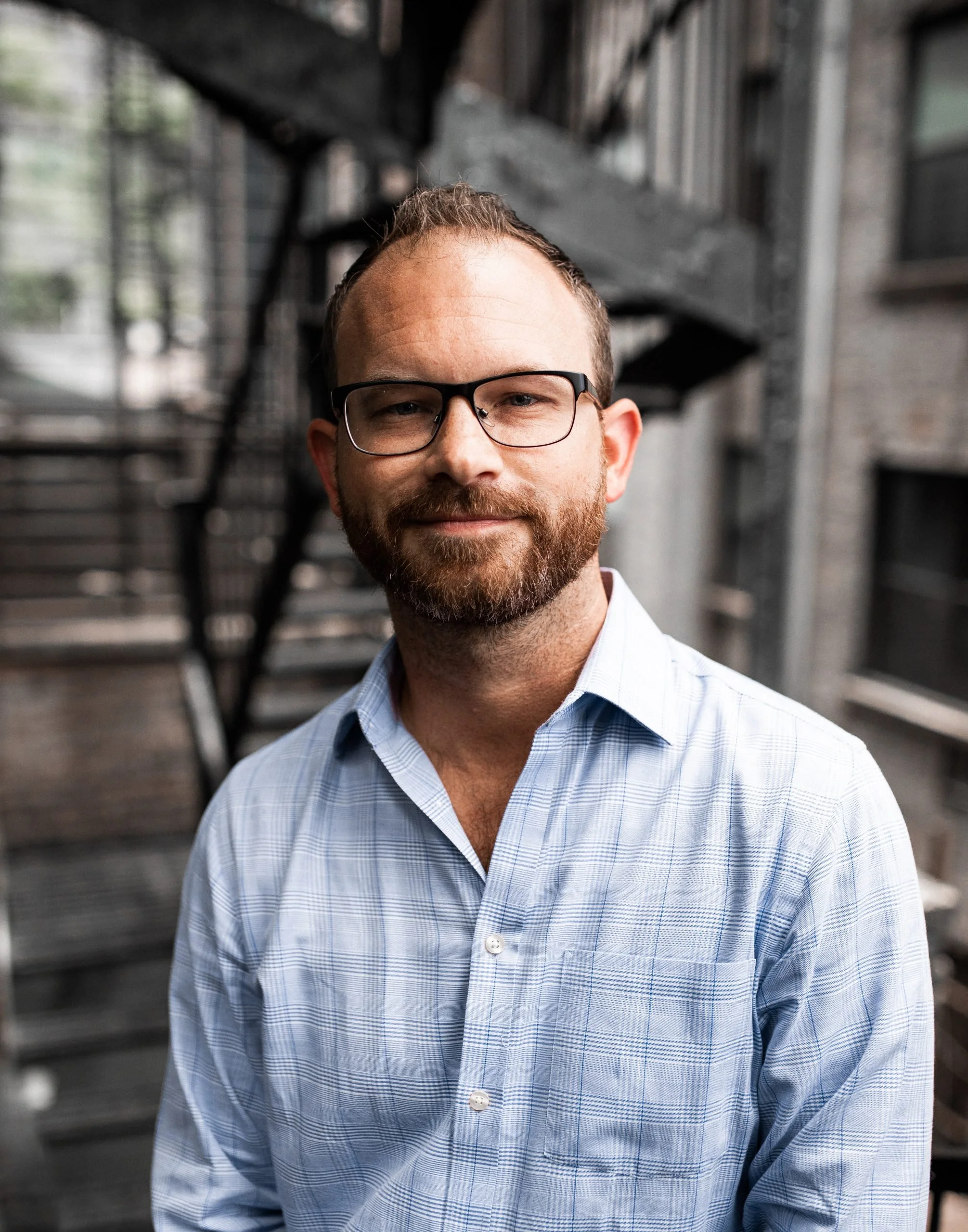 Professional man with glasses, beard, and light blue plaid shirt standing outdoors near a brick building with fire escape stairs.